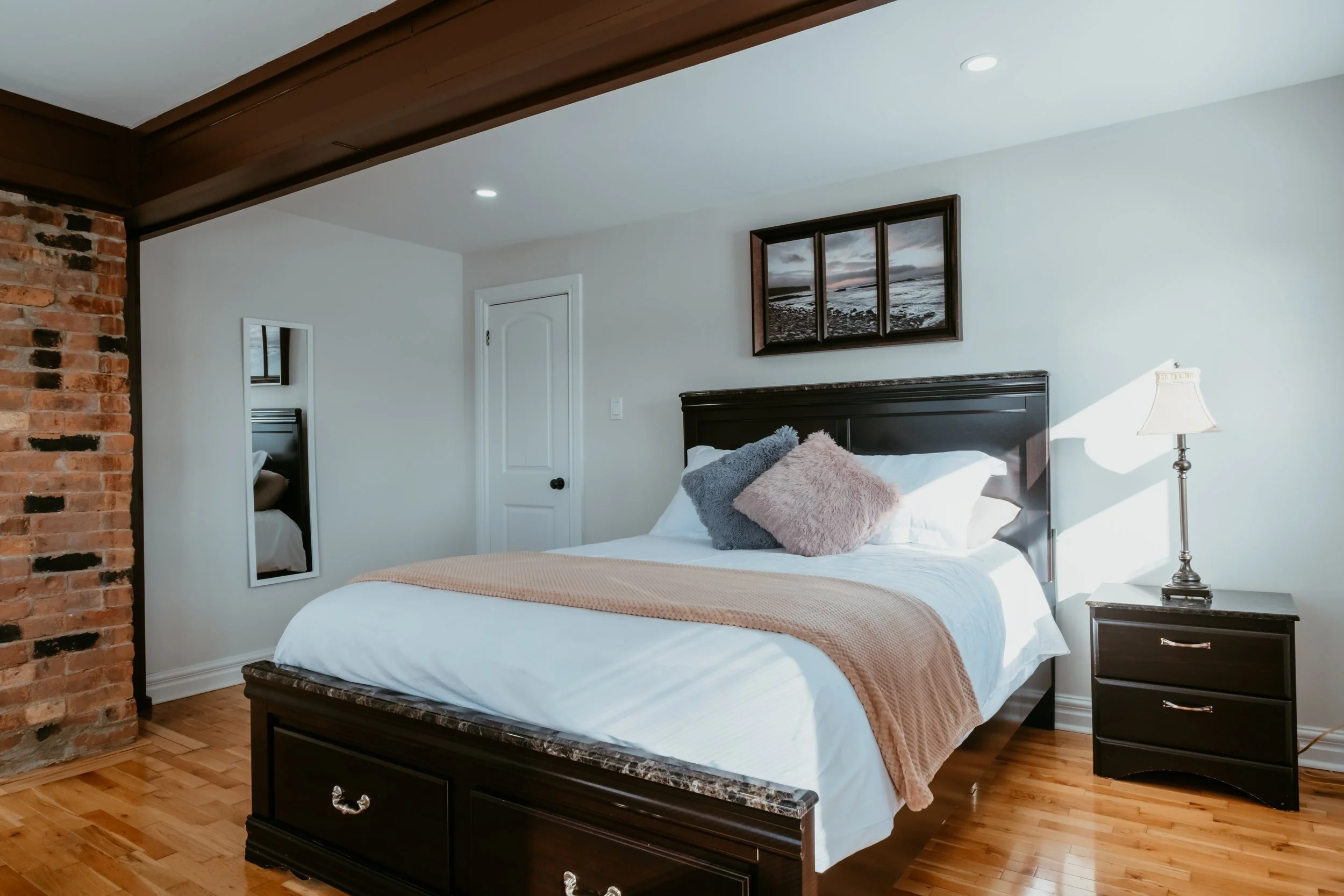 A bedroom with a bed, pillows, a nightstand, a lamp, artwork of a seascape, a mirror, and a brick column.