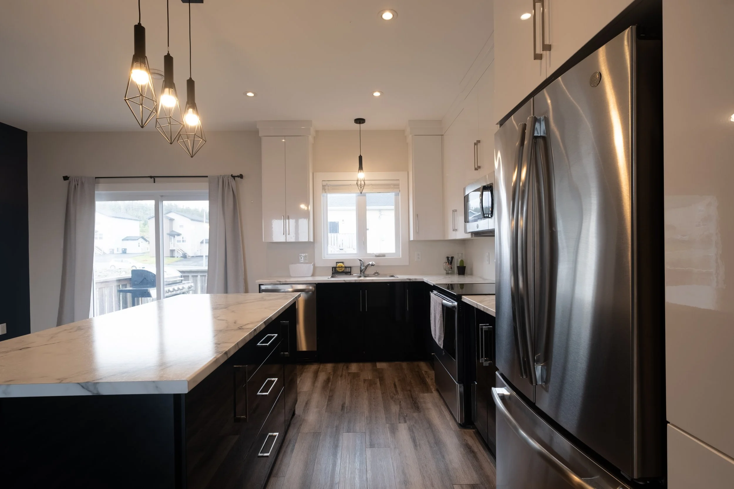 Modern kitchen with black and white cabinets, stainless steel appliances, pendant lights, and large windows with curtains.