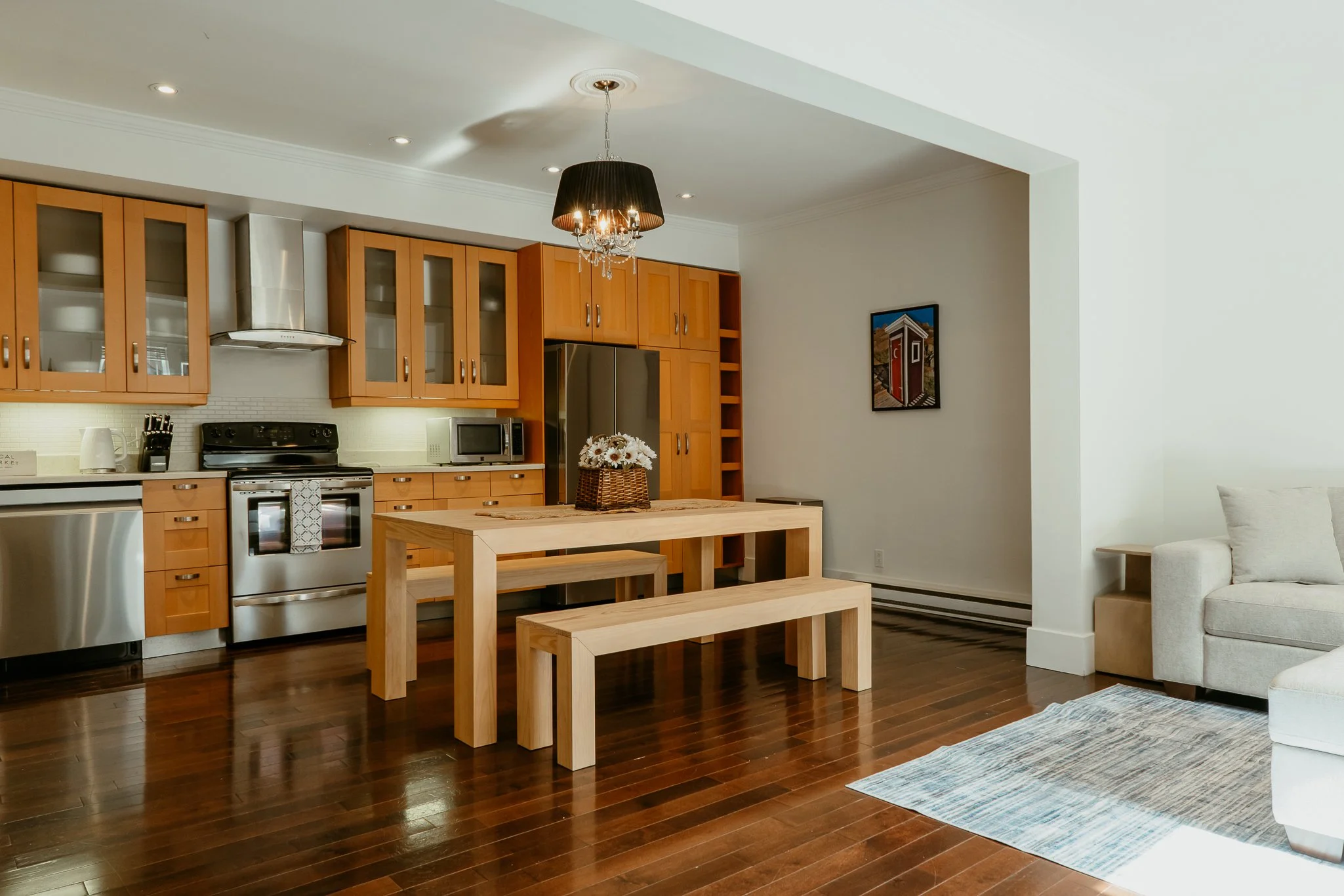Open-concept kitchen and living area with wooden floors, light-colored walls, and modern furnishings. The kitchen features wooden cabinets, stainless steel appliances, and a black pendant light with crystal accents hanging from the ceiling. A wooden table with benches and a flower basket centerpiece is in the foreground, and the living area has a beige sofa, a blue and brown area rug, and a small side table.