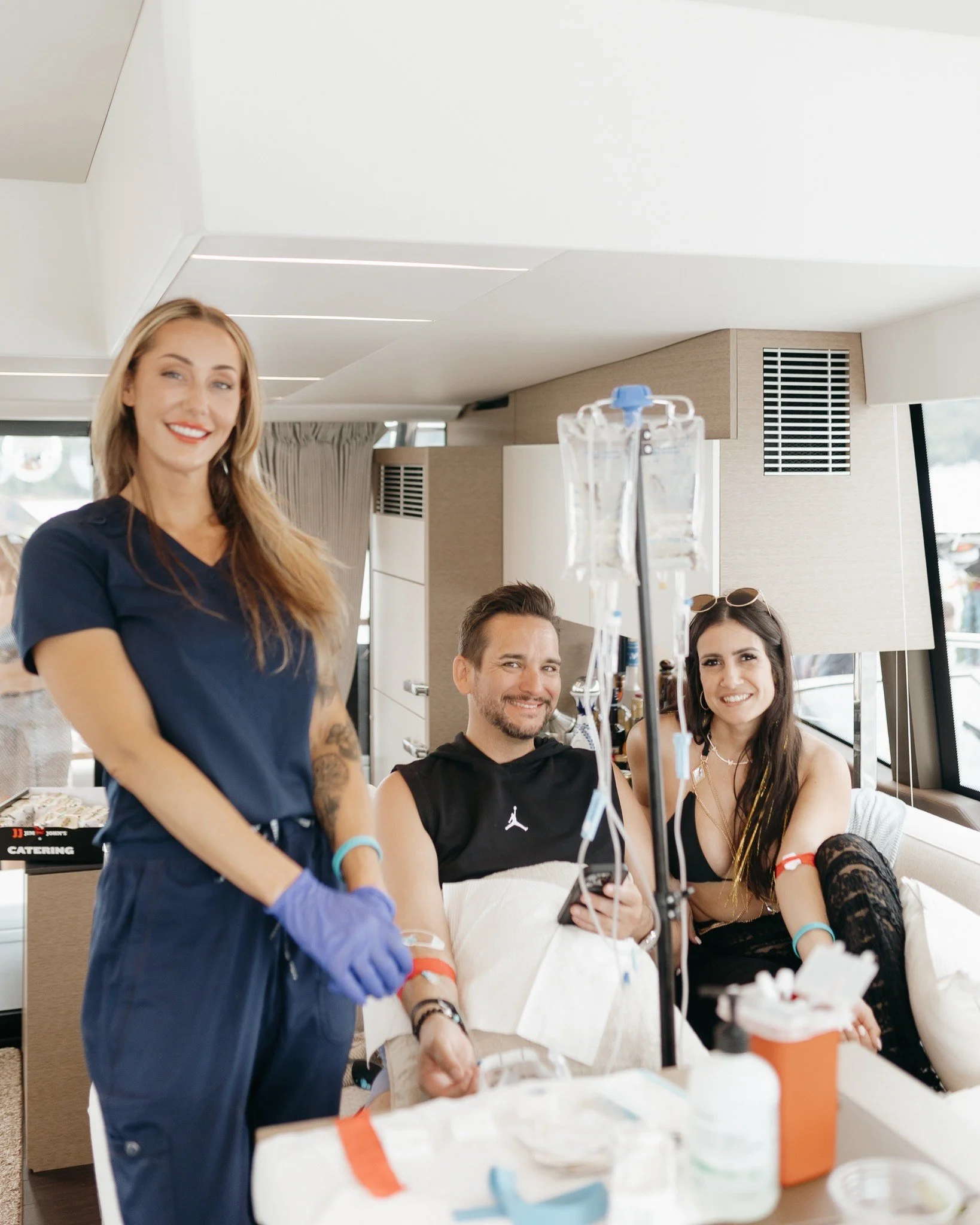 Two women and a man donating blood in a medical setting, with the woman standing in blue scrubs and gloves, and the man and woman sitting in chairs with blood donation equipment. Wellness.  Vitamin IV.  Concierge.  Mobile services.  Hangover IV.