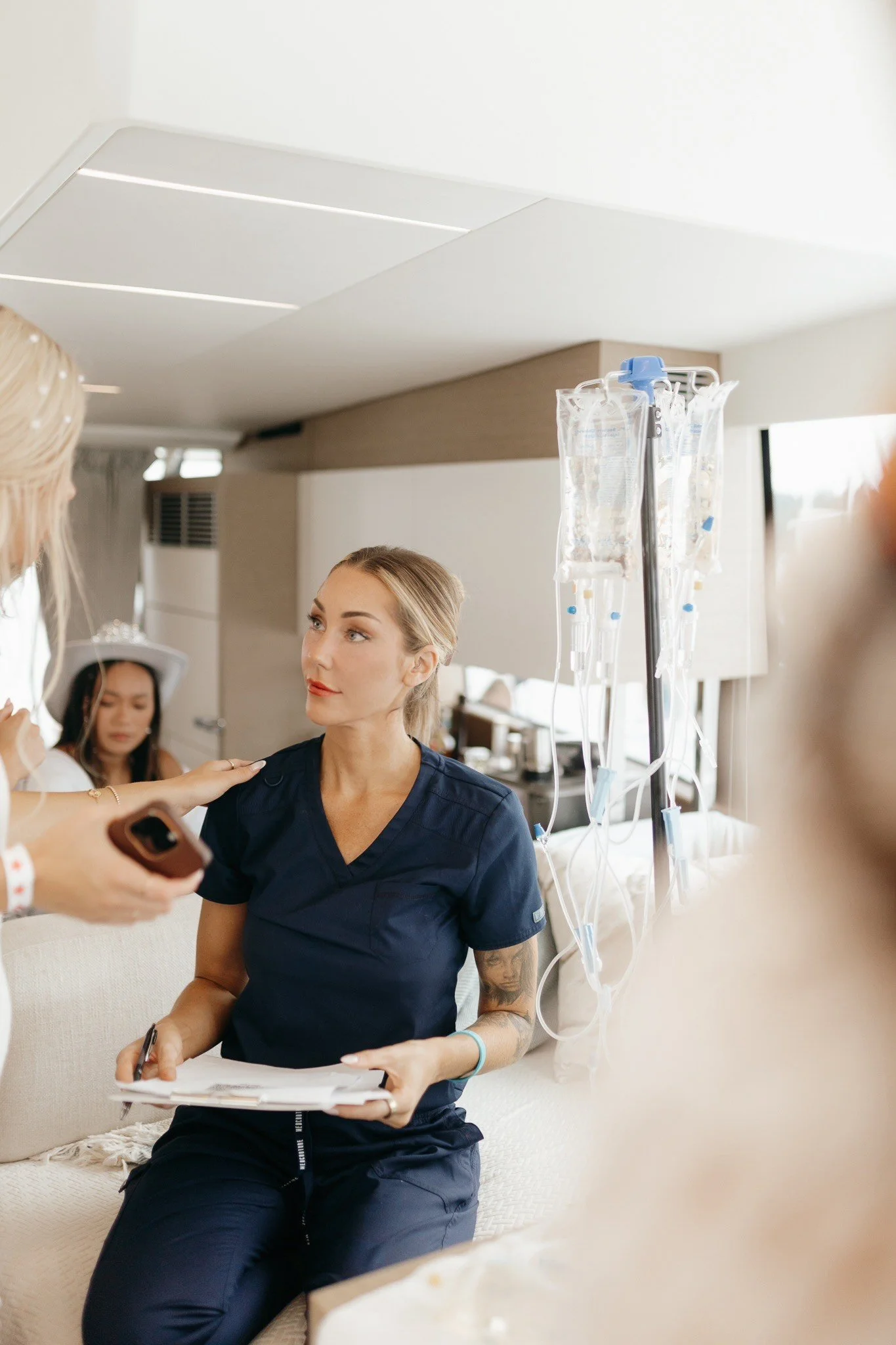 A woman in navy scrubs sitting on a beige couch with medical IV bags attached to a stand beside her, a person with a blonde ponytail and white shirt reaching out to her, and another woman in the background wearing a white hat.