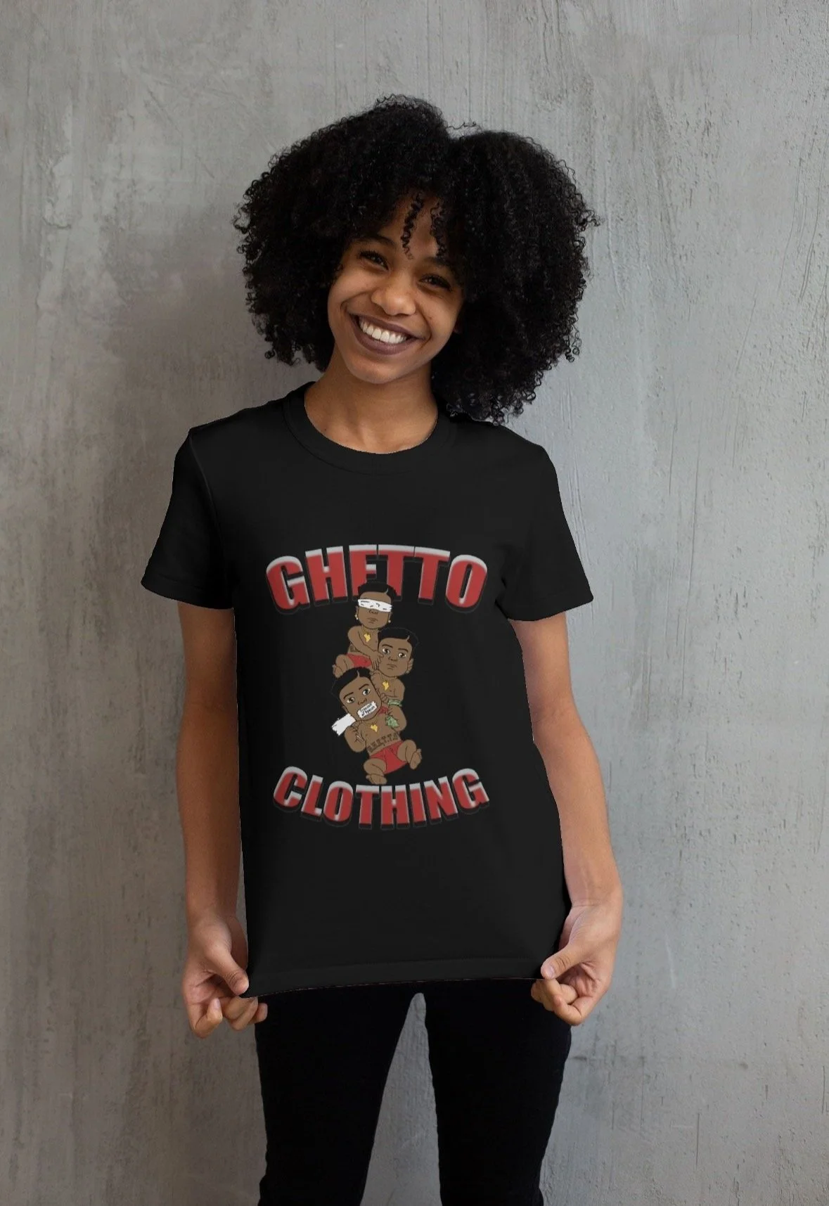 A woman with curly black hair smiling while wearing a black t-shirt with cartoon characters and the words "Ghetto Clothing" on it, standing against a plain wall.