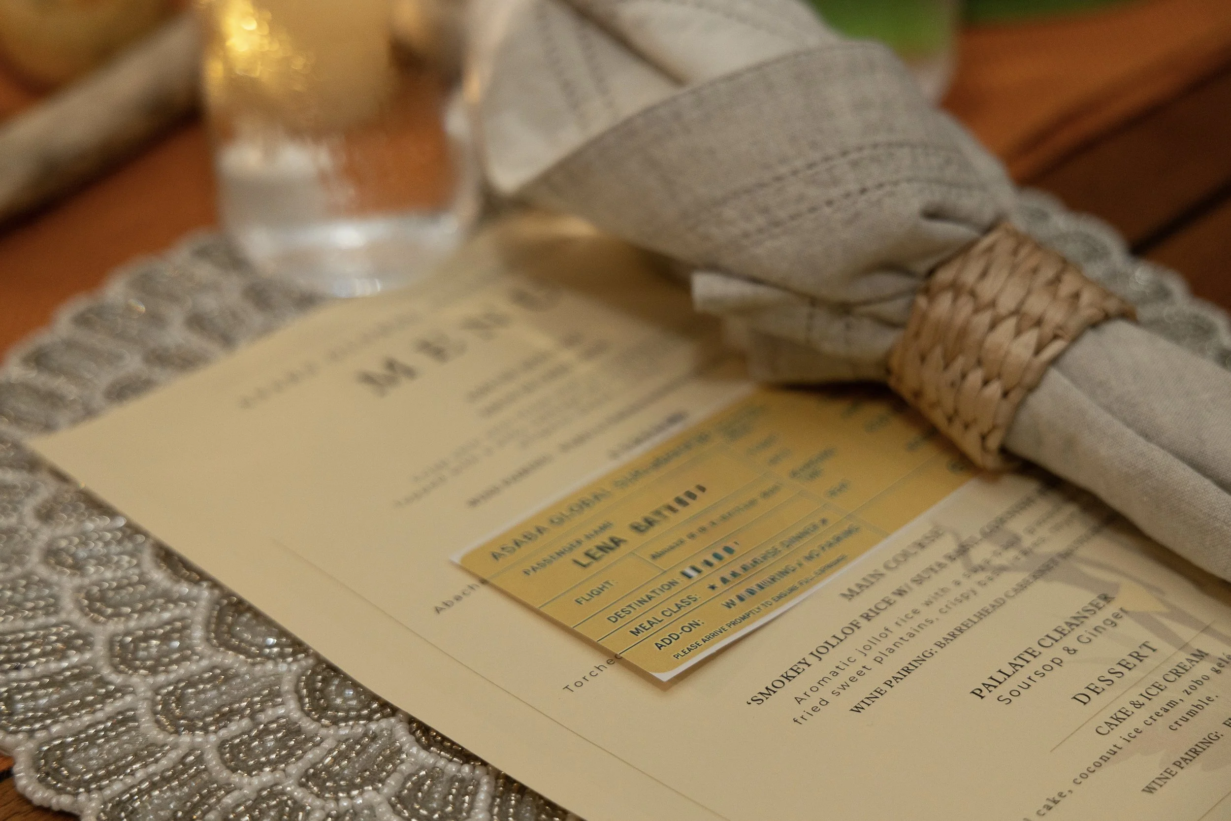 A restaurant table setting with a lace tablecloth, a folded napkin secured with a woven band, a glass of water, a menu, and a yellow boarding passt.
