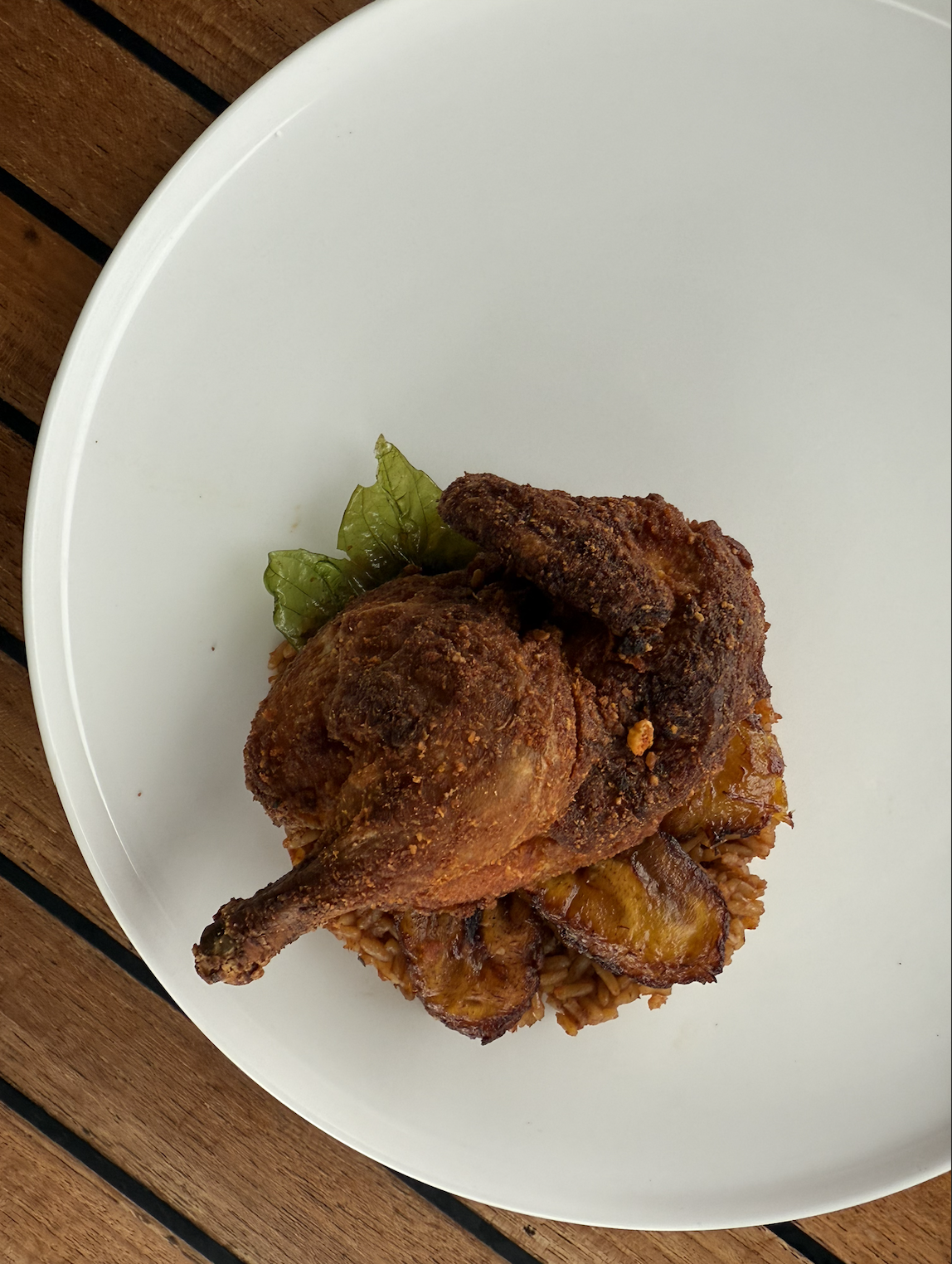 Suya Basil cornish hen with jollof rice, plantains, and garnished with a fried basil leaf.