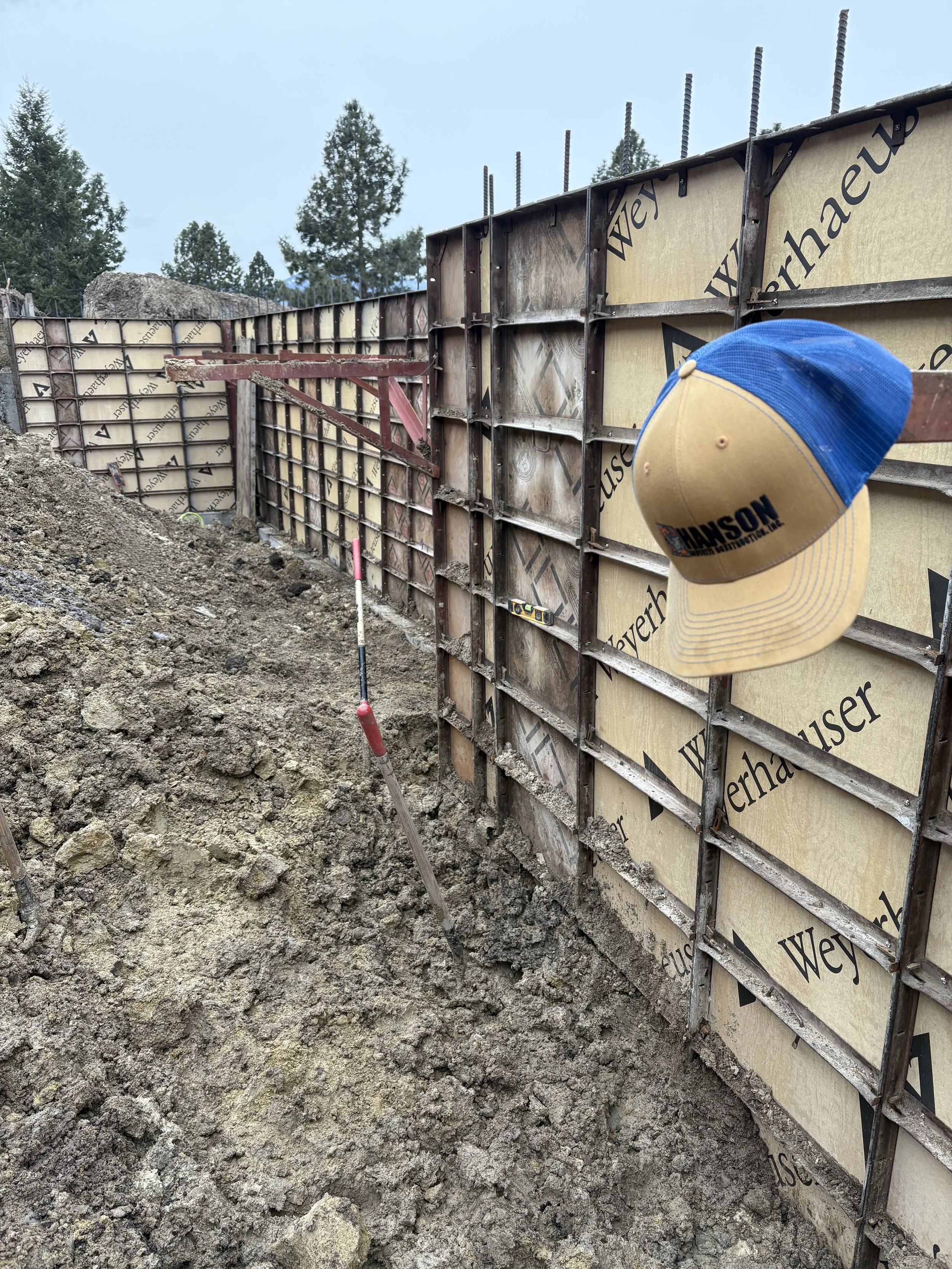 Construction site with forming walls, soil, and tools, with a blue and tan hat hanging on the wall.