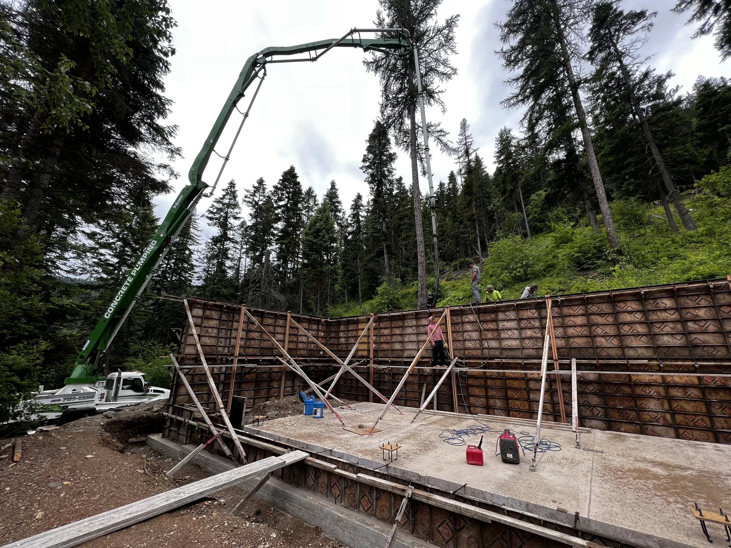 Construction workers are building a concrete wall in a forested area, using a large concrete pump truck to pour concrete.