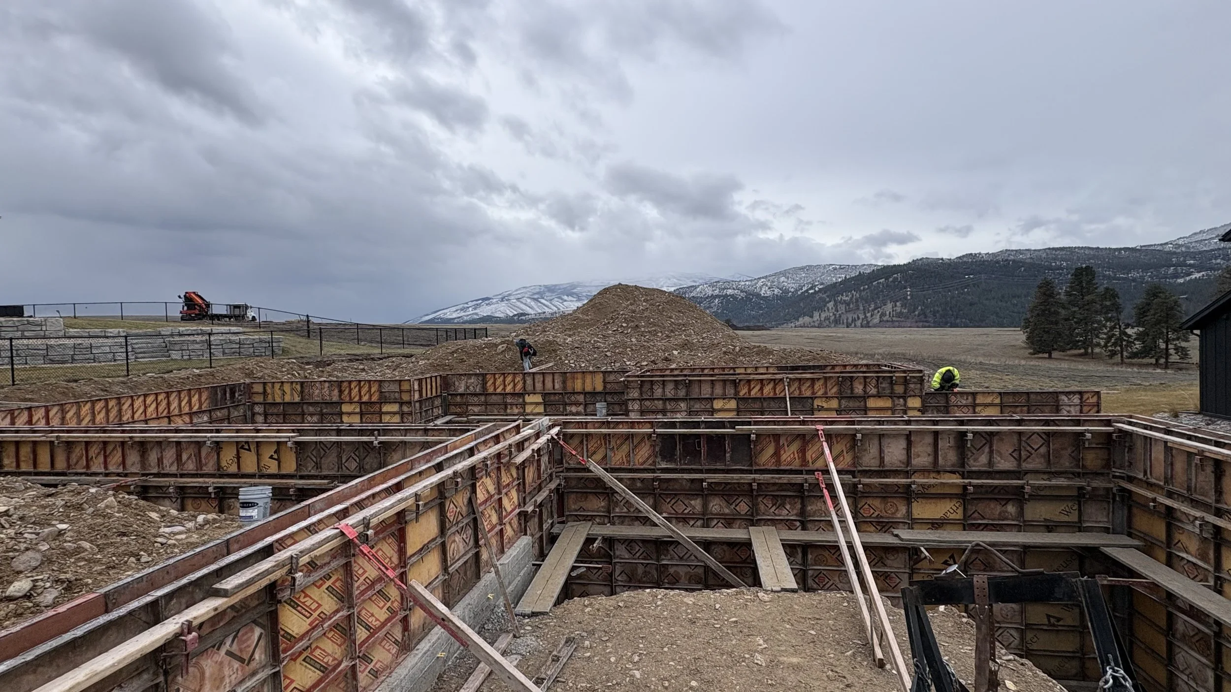 Construction site with workers building a foundation, surrounded by a rural landscape with mountains and cloudy sky.