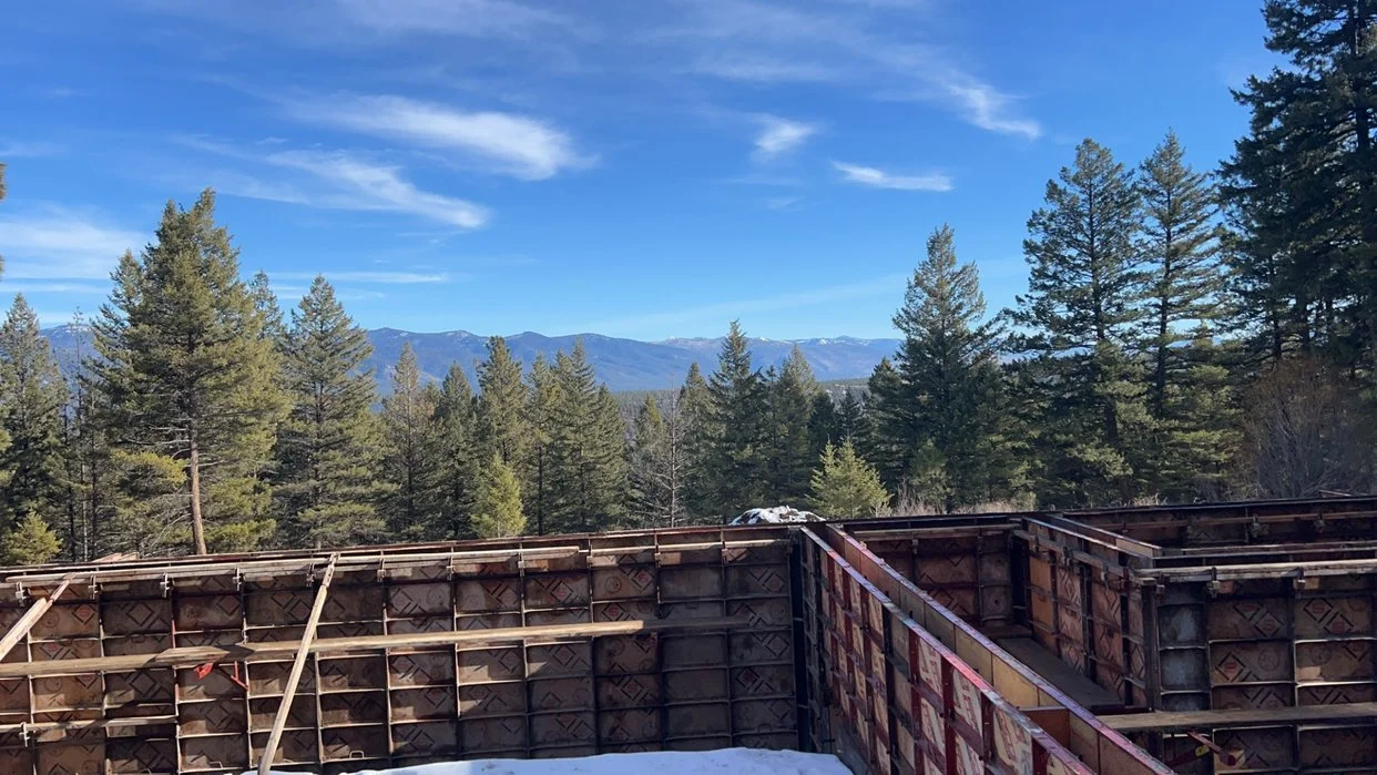 View of a construction site with concrete formwork, surrounded by tall evergreen trees and distant mountains under a blue sky.