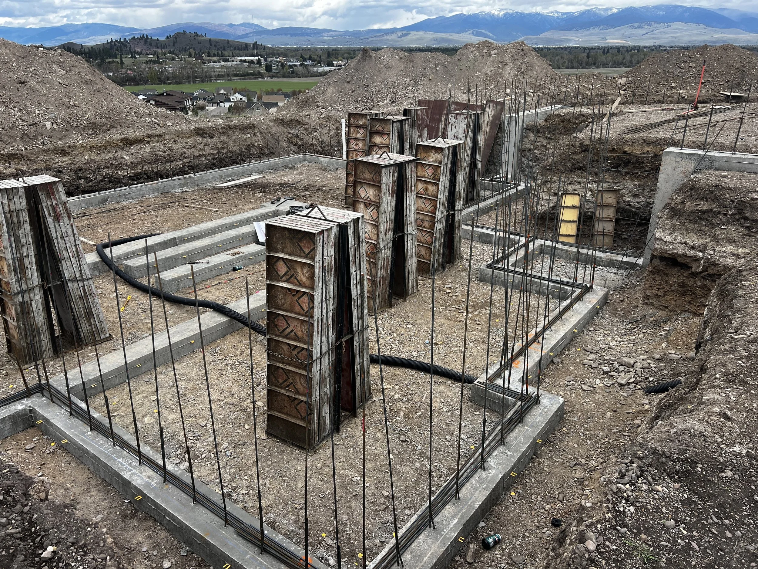 Construction site showing the foundation for a building with metal reinforcement bars and concrete forms in place, overlooking a landscape of hills and a cloudy sky.