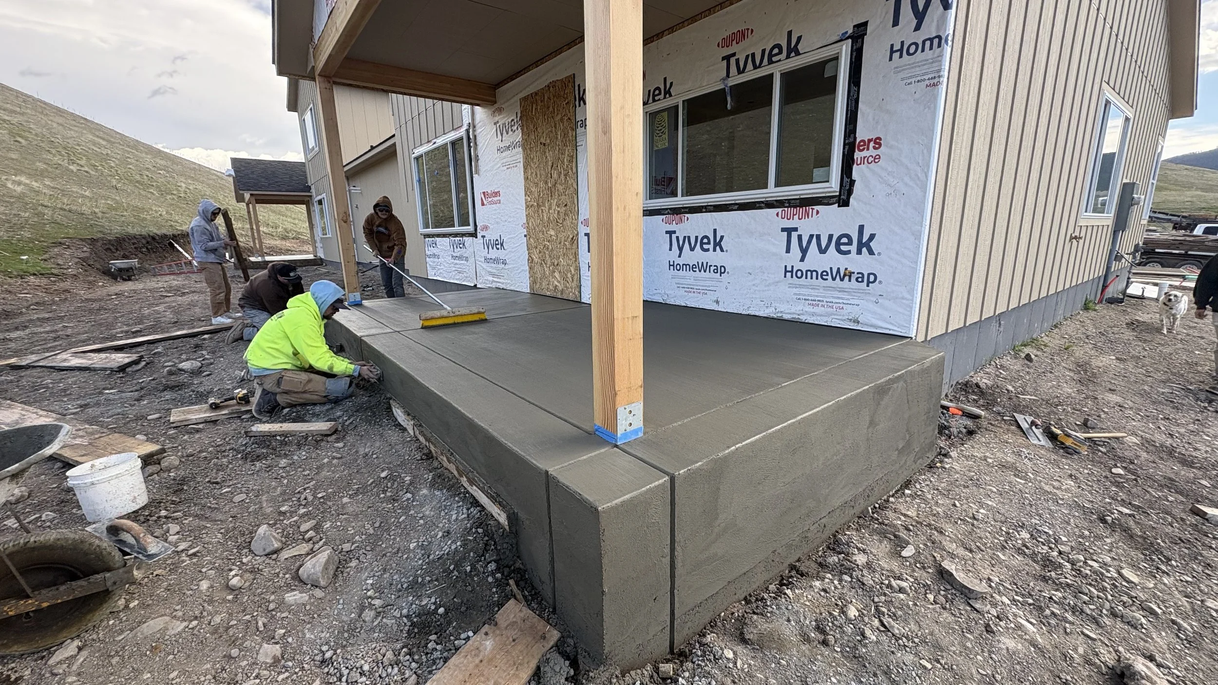 Construction workers are pouring and smoothing a concrete porch in front of a house under construction, with some workers kneeling and others standing, using tools, while a dog is in the background near the house.