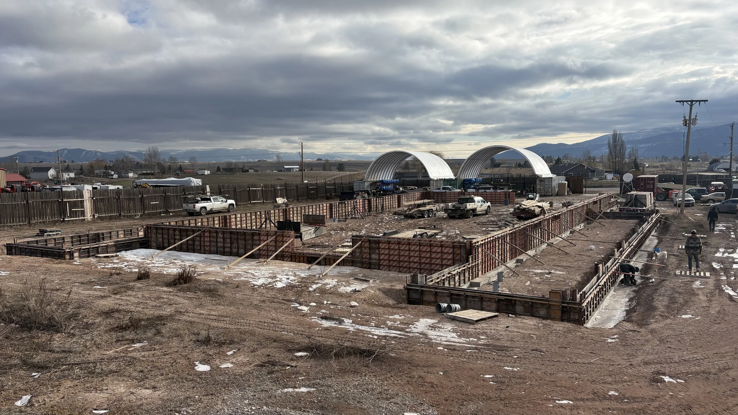 Construction site with concrete foundation being prepared, surrounded by wooden frames, with workers working, stationary trucks, and vehicles behind, under a cloudy sky with mountains in the distance.