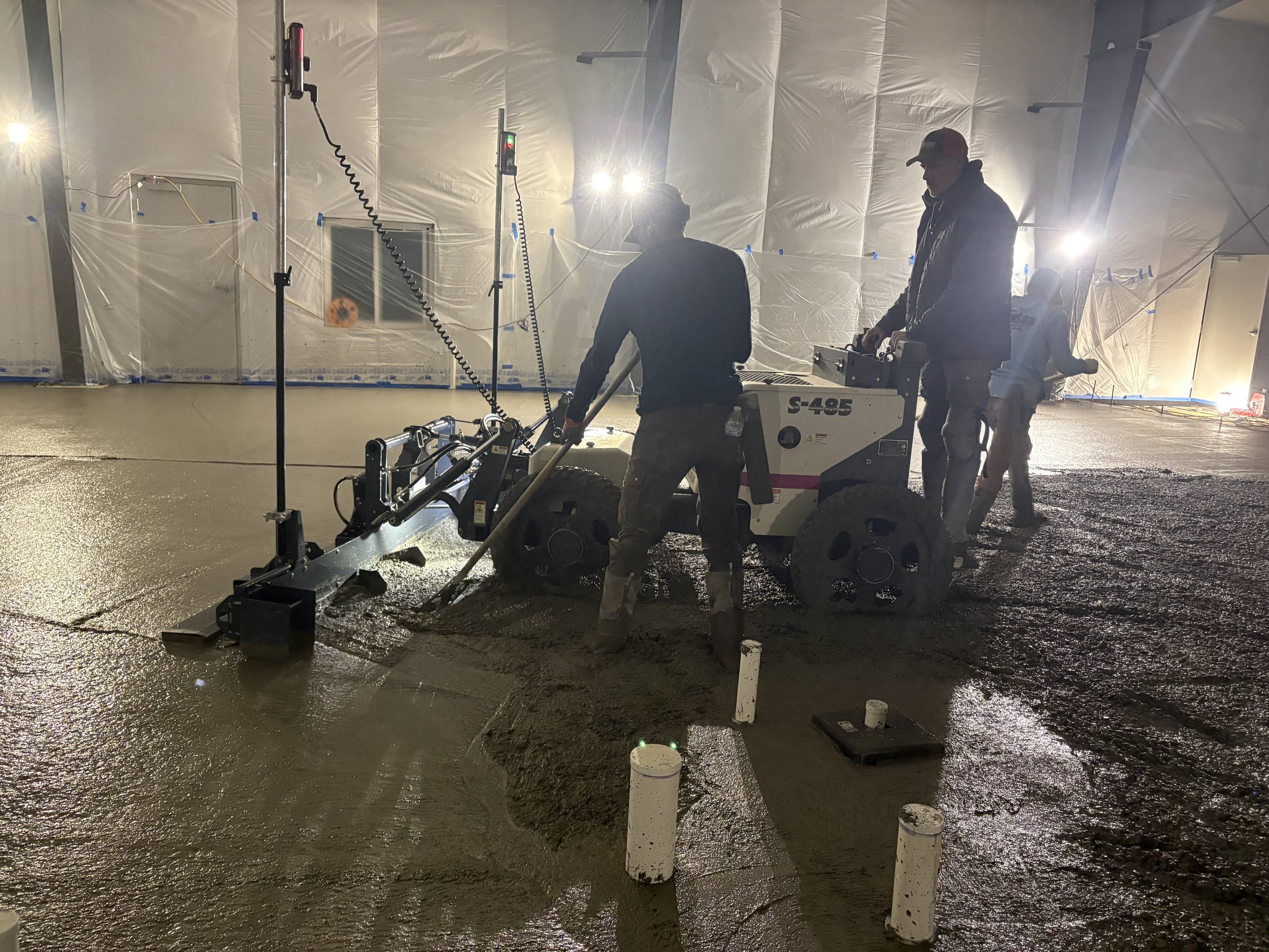 Workers operating machinery on freshly poured concrete floor in an indoor construction site with plastic sheeting covering walls and bright lighting.
