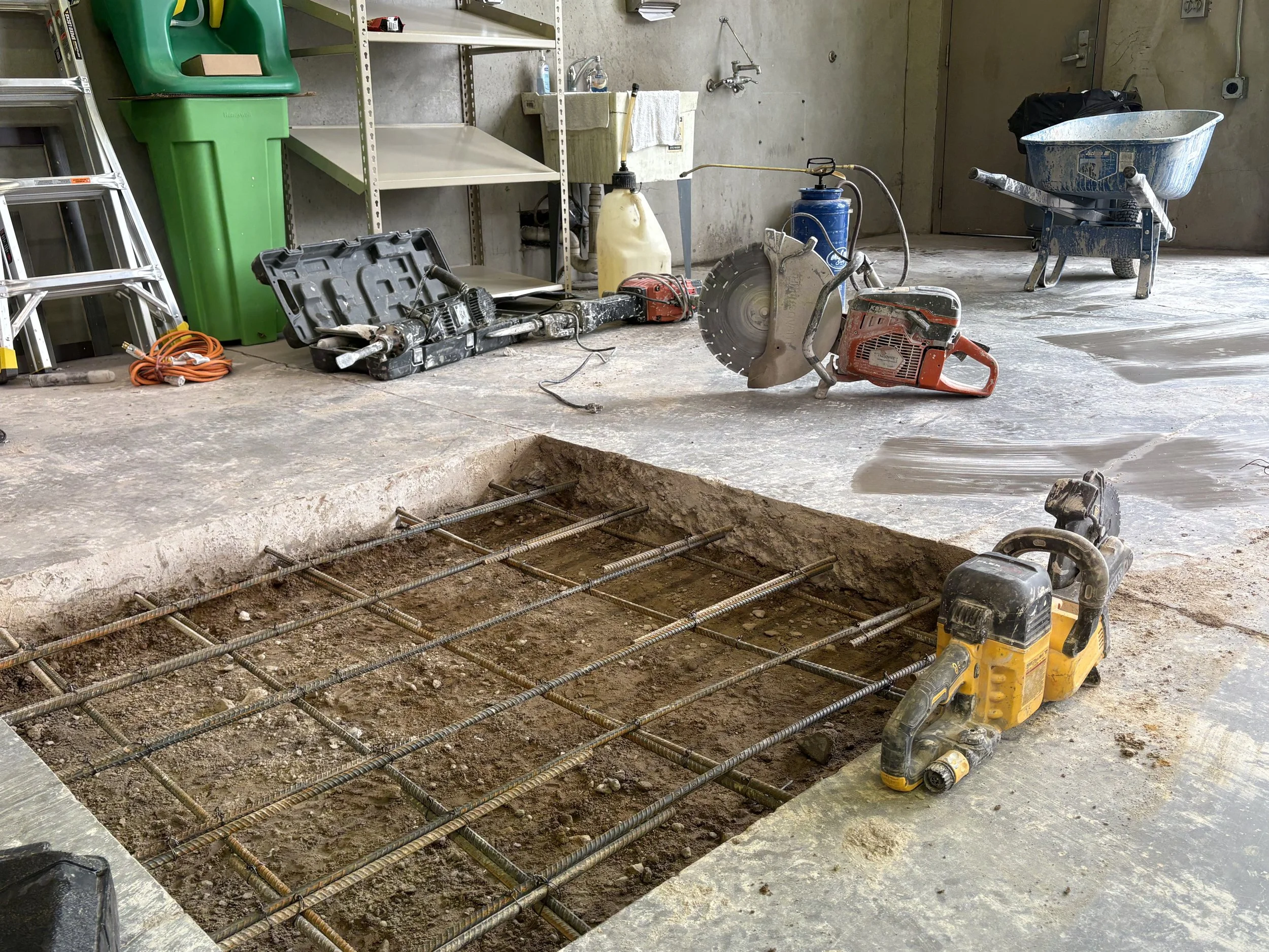 Construction site with rebar grid for concrete pouring, construction tools and equipment including a saw, drill, and water hose, in an indoor space with shelves, a wheelbarrow, and concrete walls.