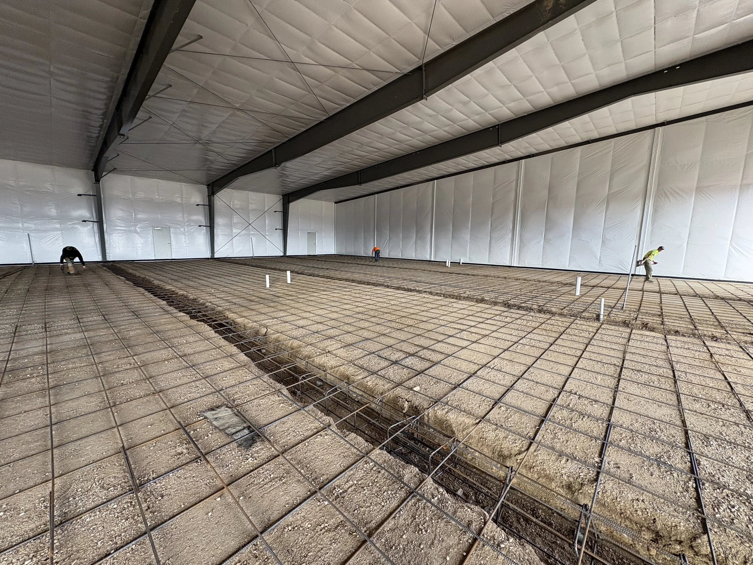 Workers are installing a concrete floor over a wire mesh framework inside a large warehouse-like building with insulated walls and a high ceiling.