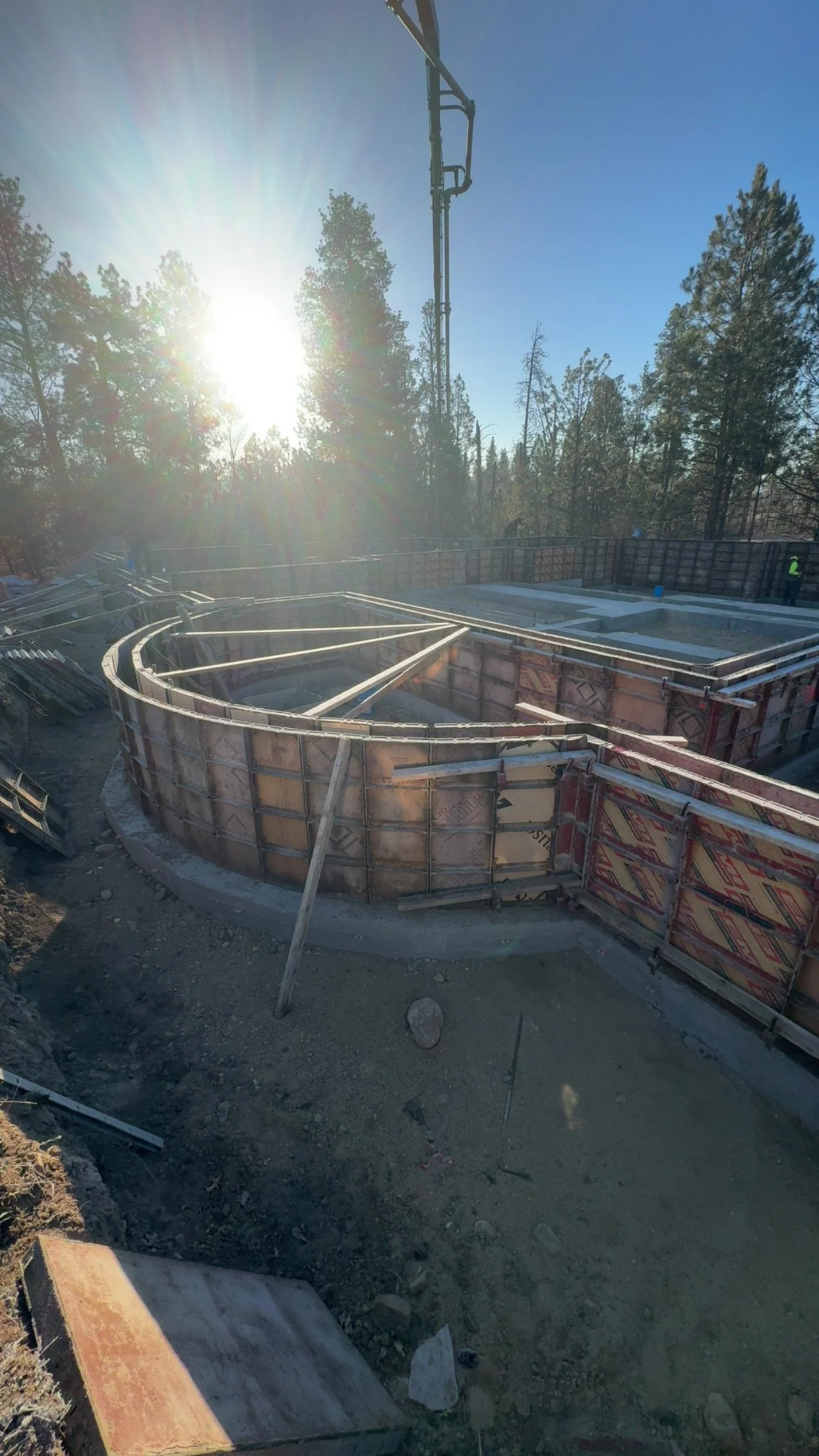 Construction site with a curved concrete foundation and wooden formwork, surrounded by trees and sunlight.