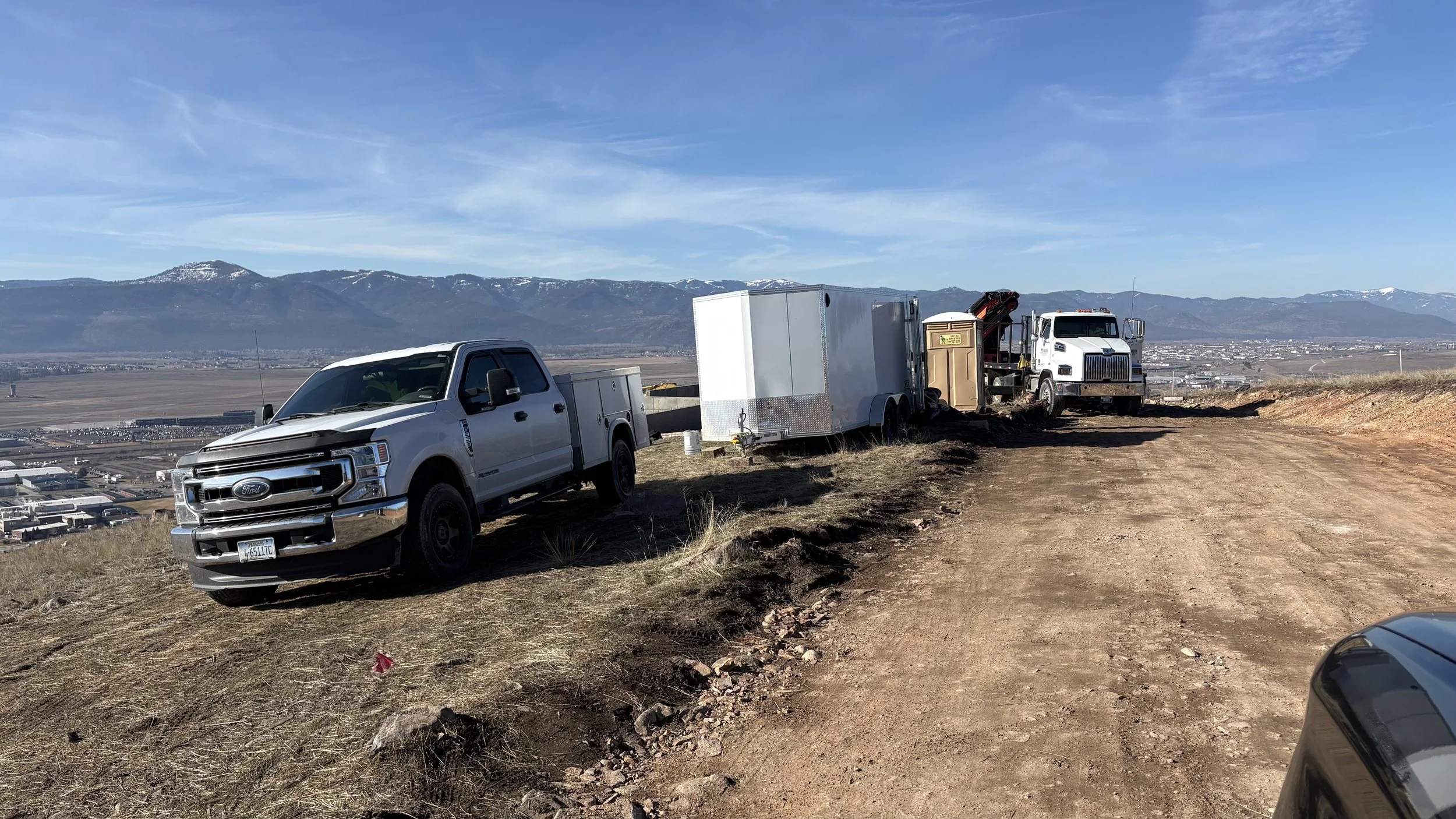 Construction vehicles and trucks on a dirt hill top with a mountain and city in the distance under a blue sky.