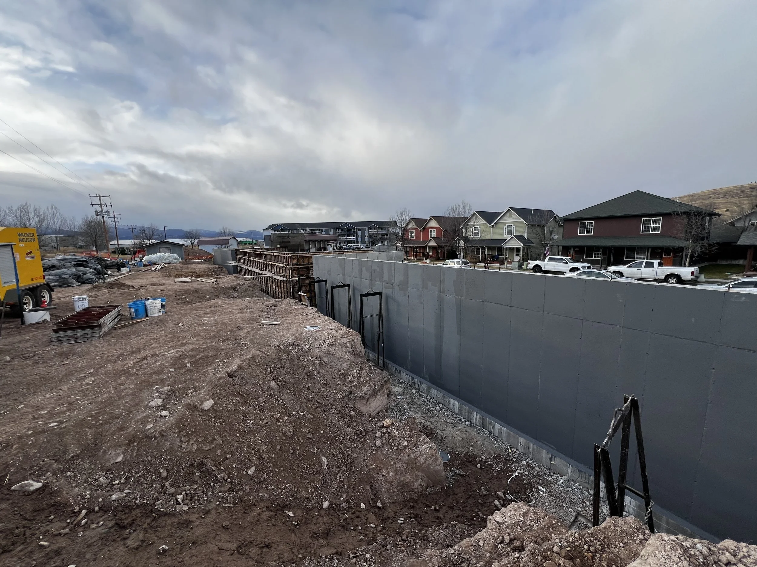 Construction site with dirt and gravel foreground, and a partially built wall with wooden supports in the middle ground, with residential houses and parked cars in the background under a cloudy sky.