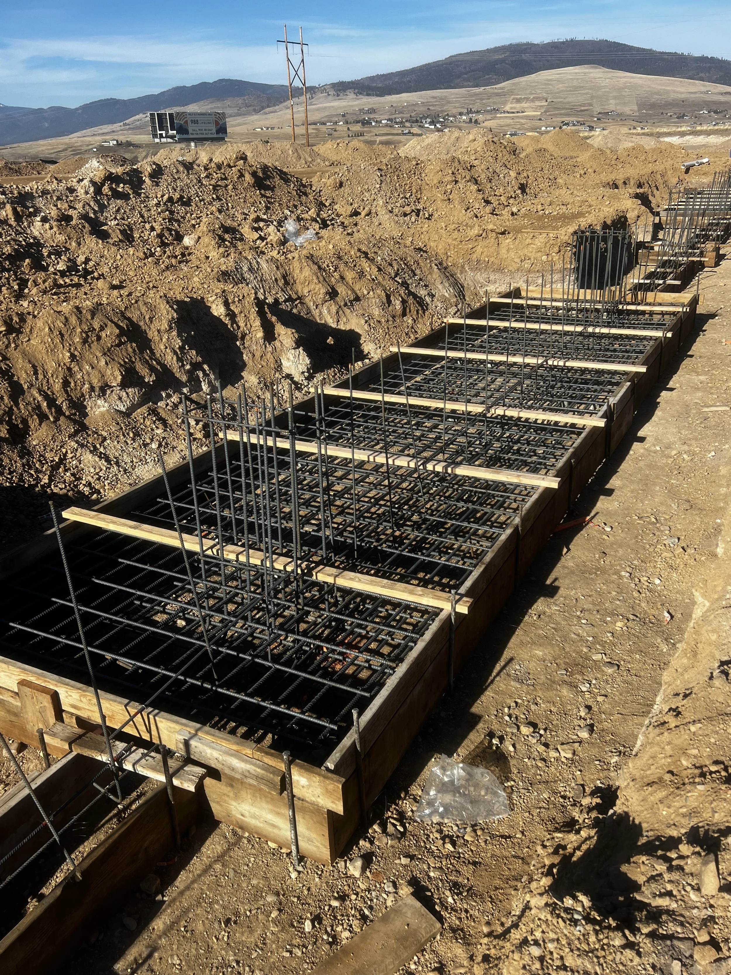 Construction site with rebar reinforced concrete formwork in a dirt trench, with hills and a clear blue sky in the background.