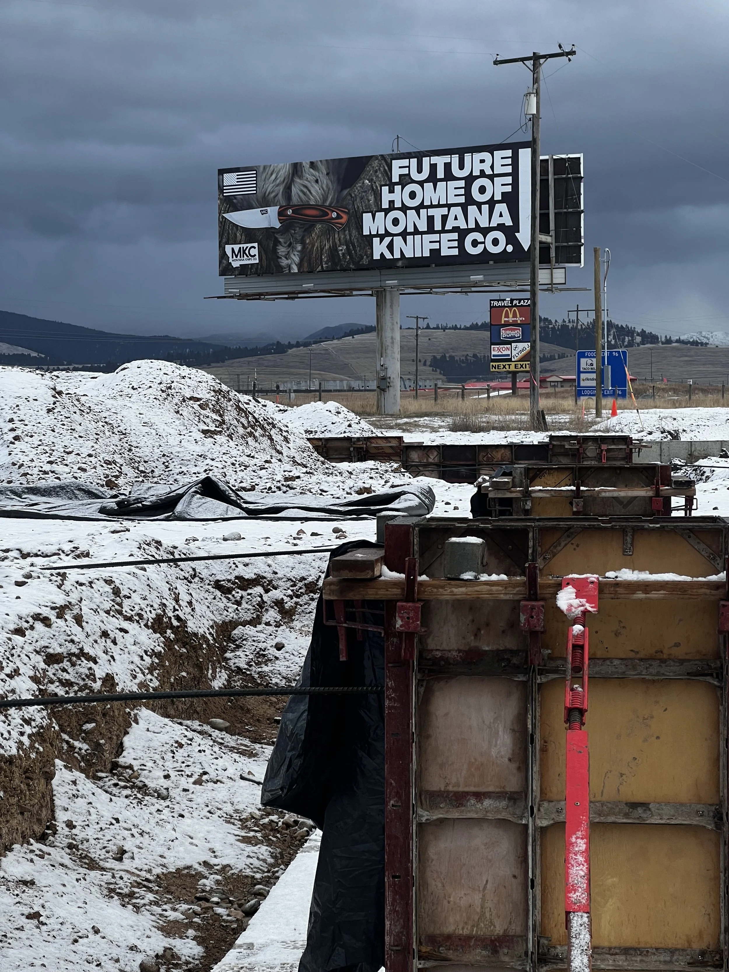 A billboard with the message 'Future home of Montana Knife Co.' and an image of a knife, set against a cloudy sky in a construction area with snow on the ground.