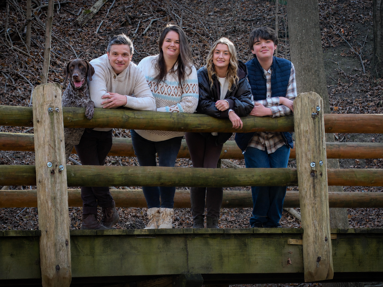Four people and a dog leaning on a wooden fence in a wooded area outdoors.