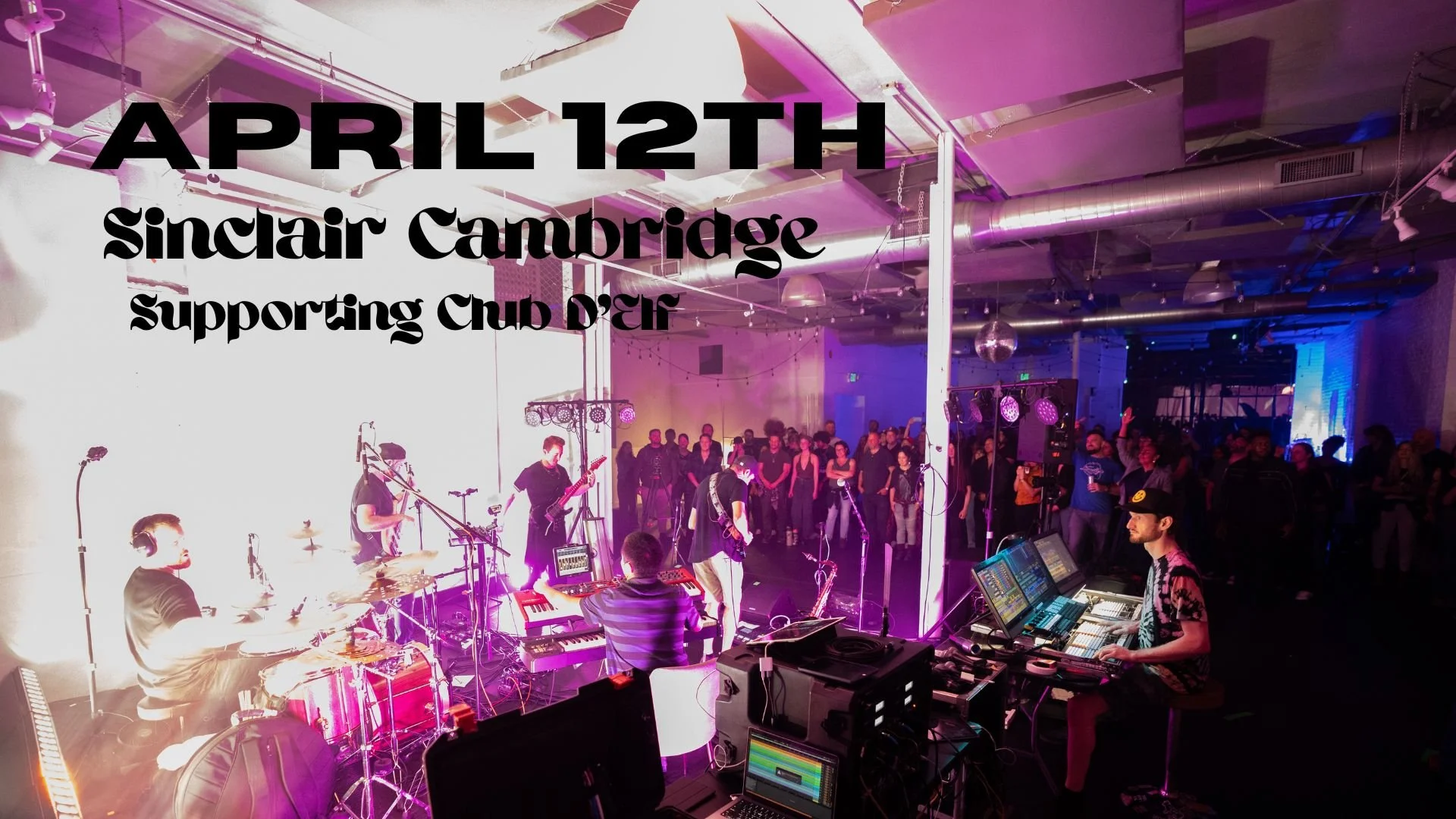 A band performs on stage at an indoor concert event, with the audience watching. Overhead lighting and equipment are visible, and a large crowd is gathered in front of the stage.