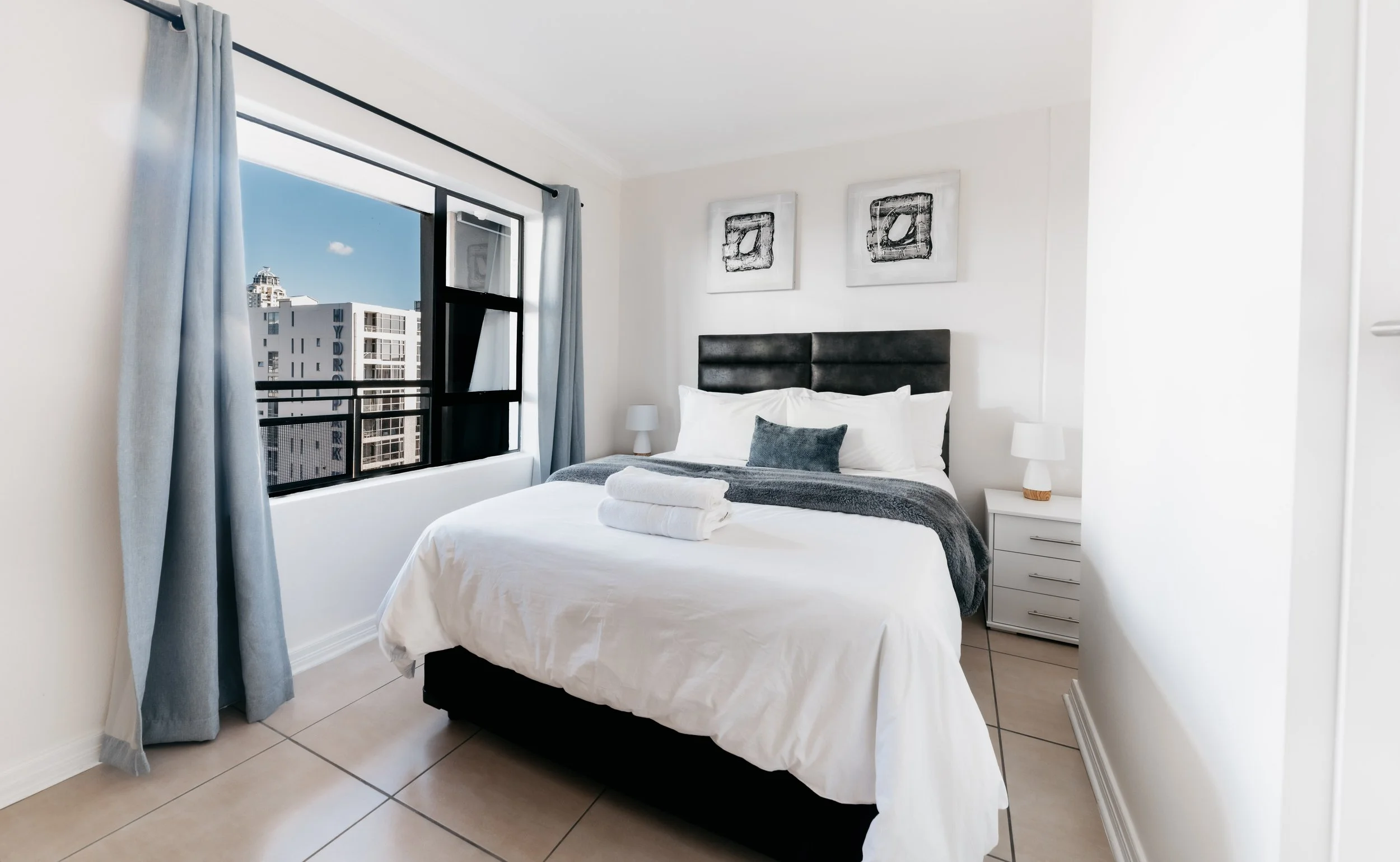 Clean, modern hotel bedroom with a large window, white walls, dark headboard, white bedding, gray pillows and blanket, and a rolled towel on the bed.