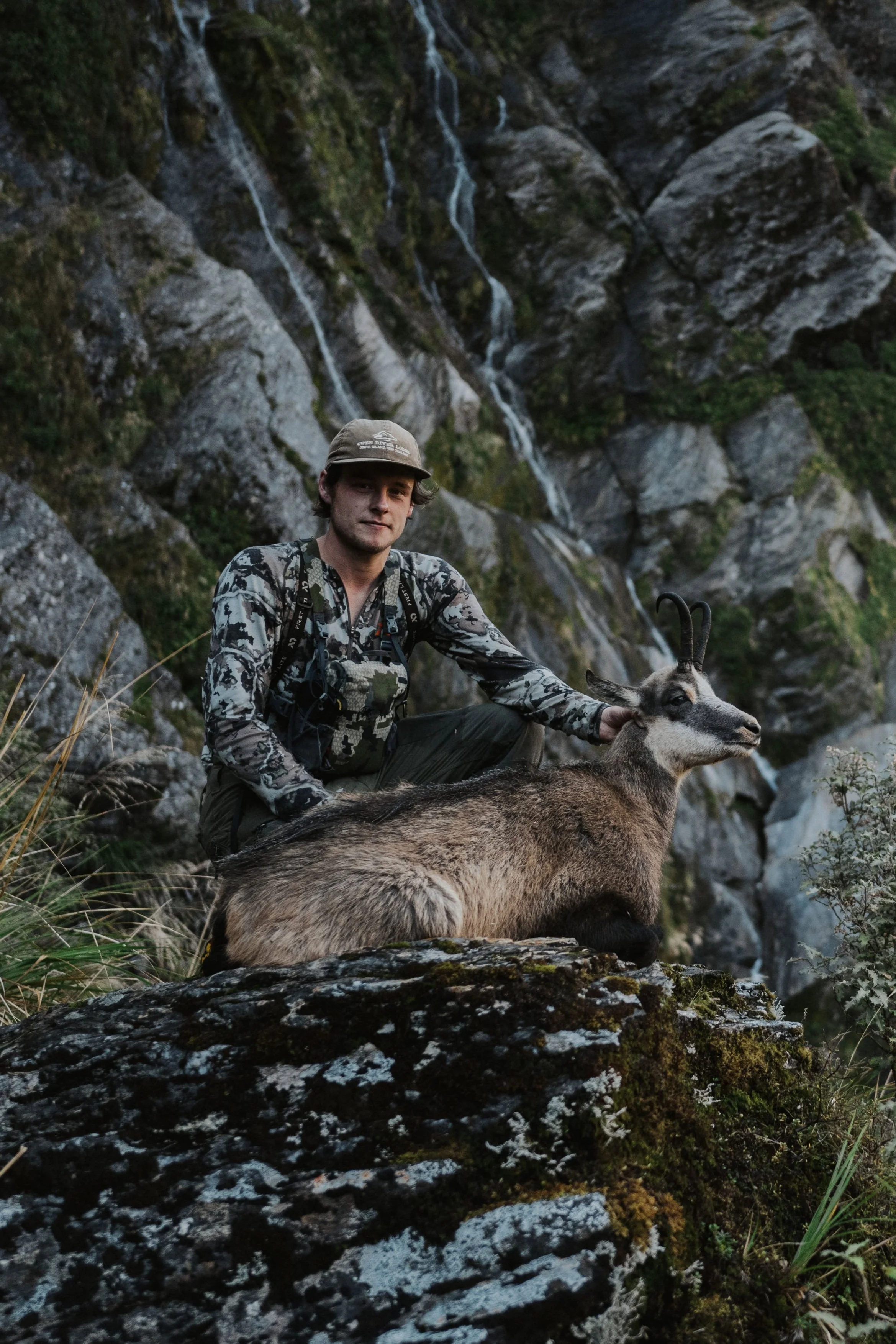 A man in outdoor camouflage clothing kneels next to a mountain goat with curved horns, in front of a rocky landscape with a waterfall.