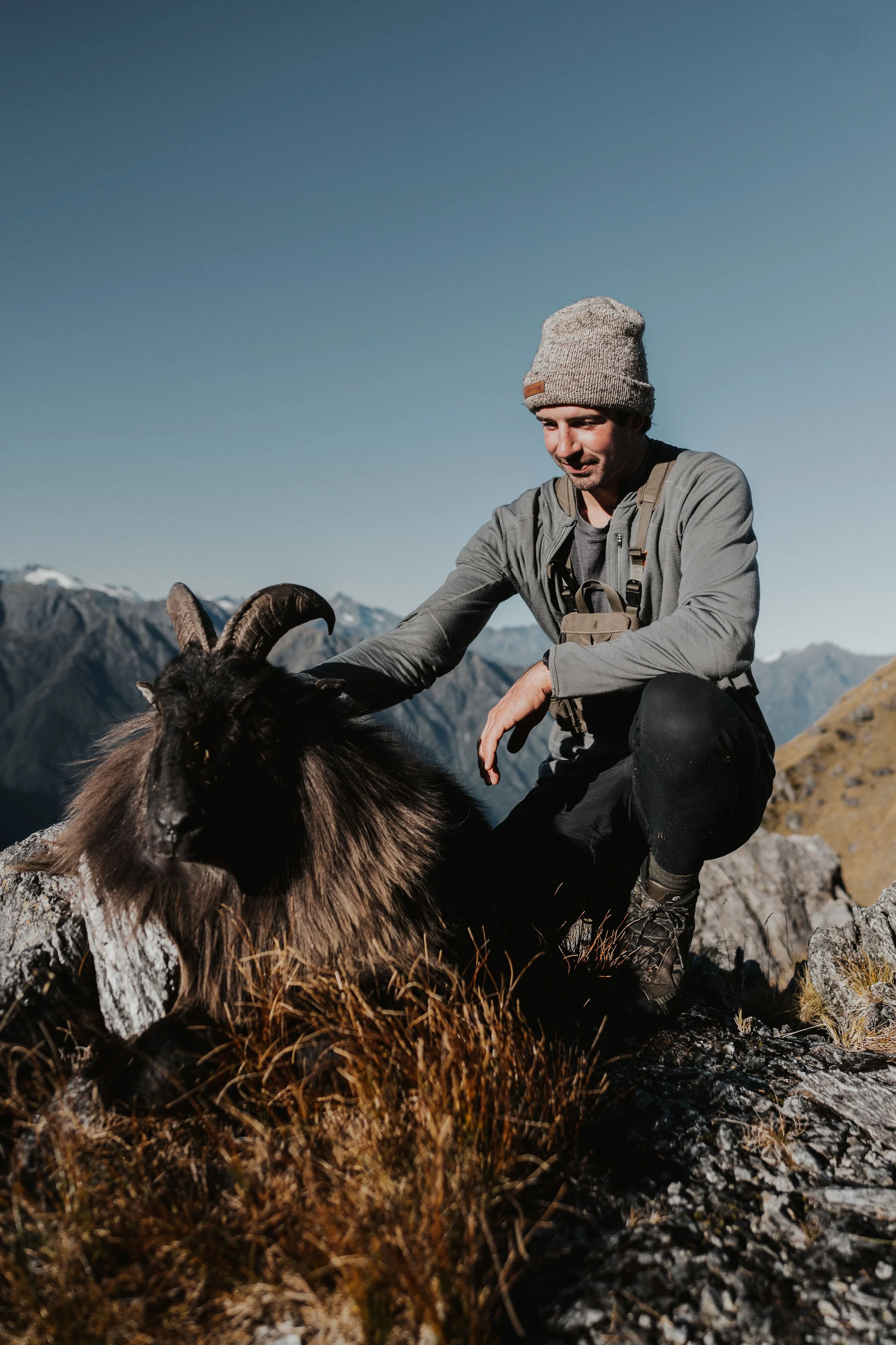 A man wearing outdoor gear, including a beanie and hiking boots, kneels next to a mountain goat with large curved horns on rocky terrain in a mountainous landscape.