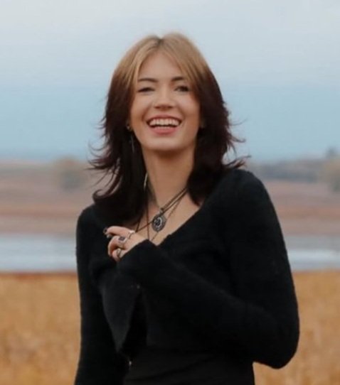 A young woman with shoulder-length brown hair smiling outdoors near a body of water and open landscape.