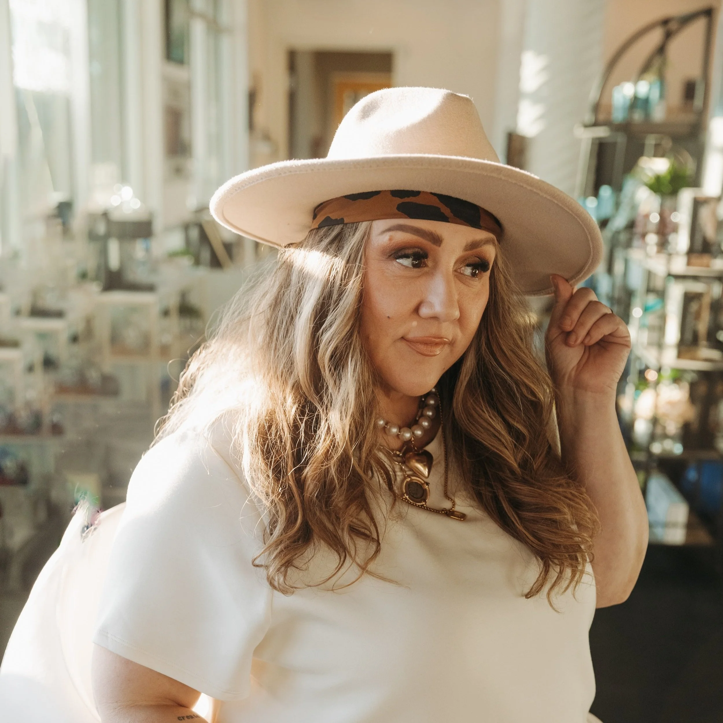 Woman with long wavy hair wearing a beige wide-brimmed hat with an animal print band, pearl necklace, and white top in a well-lit room with shelves and pictures in the background.