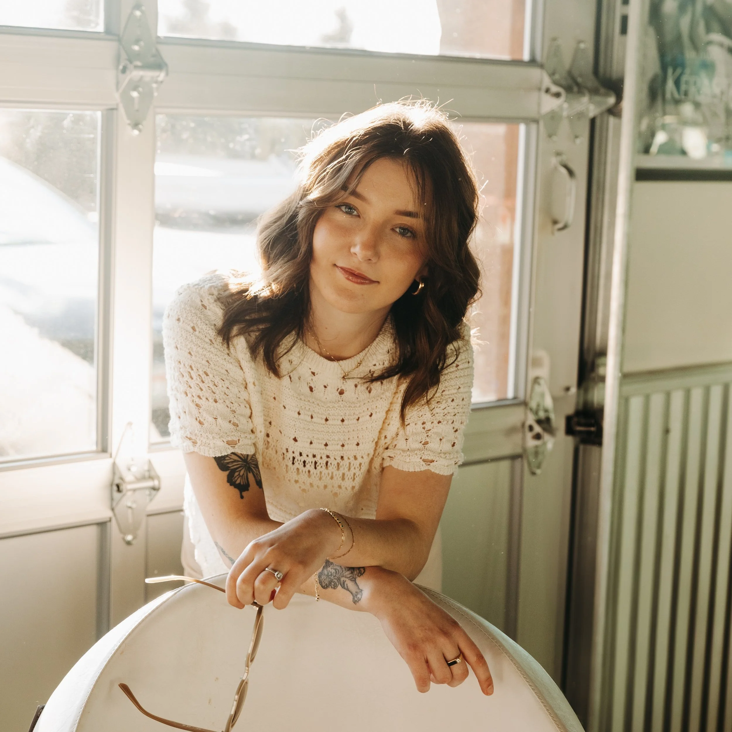 Young woman with wavy brown hair and tattoos, wearing a cream crochet top, sitting by a window with sunlight, holding sunglasses on a table inside a cafe or diner.