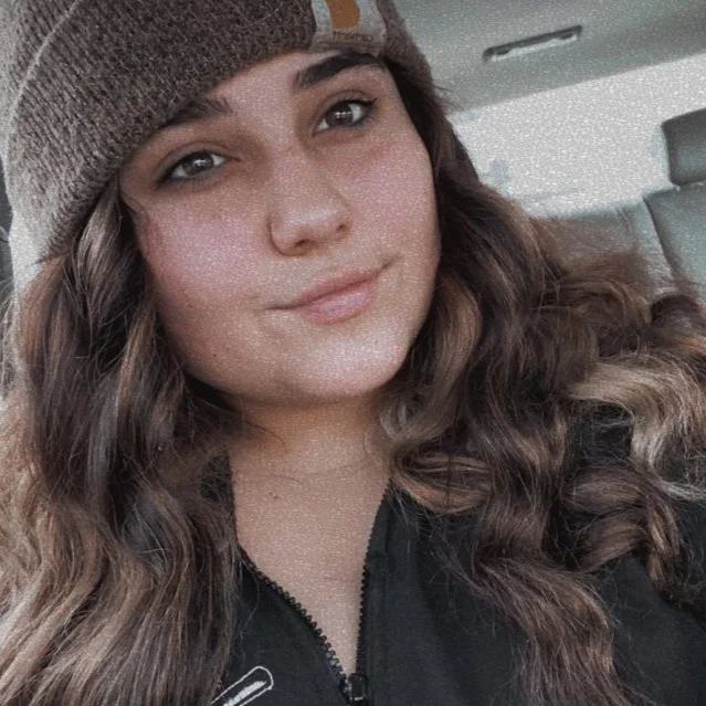 Young woman with long wavy brown hair, wearing a knit beanie and a zip-up jacket, sitting inside a vehicle.