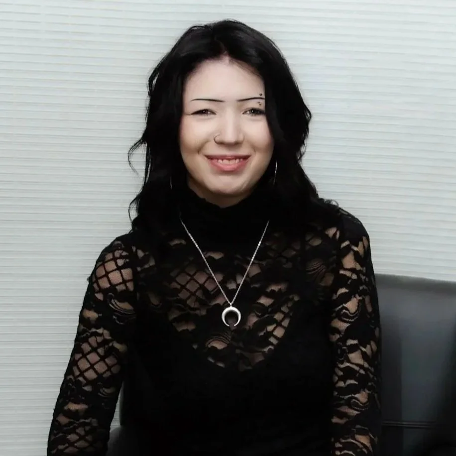A young woman with shoulder-length brown hair smiling outdoors near a body of water and open landscape.