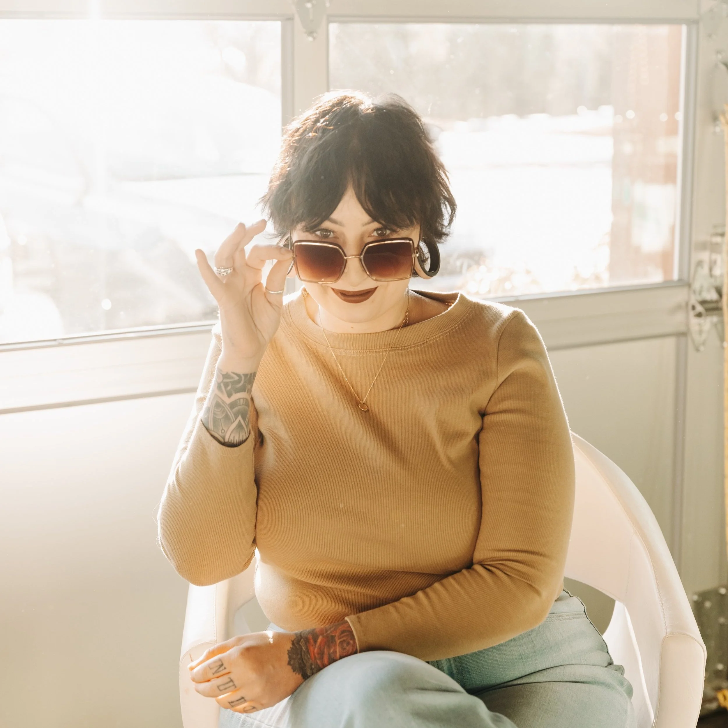 A woman with dark, curly hair and tattoos on her wrist sits in a bright room with sunlight streaming through a window. She wears a beige long-sleeve top, large sunglasses, and dark lipstick, and looks playfully at the camera while holding her sunglasses with one hand.