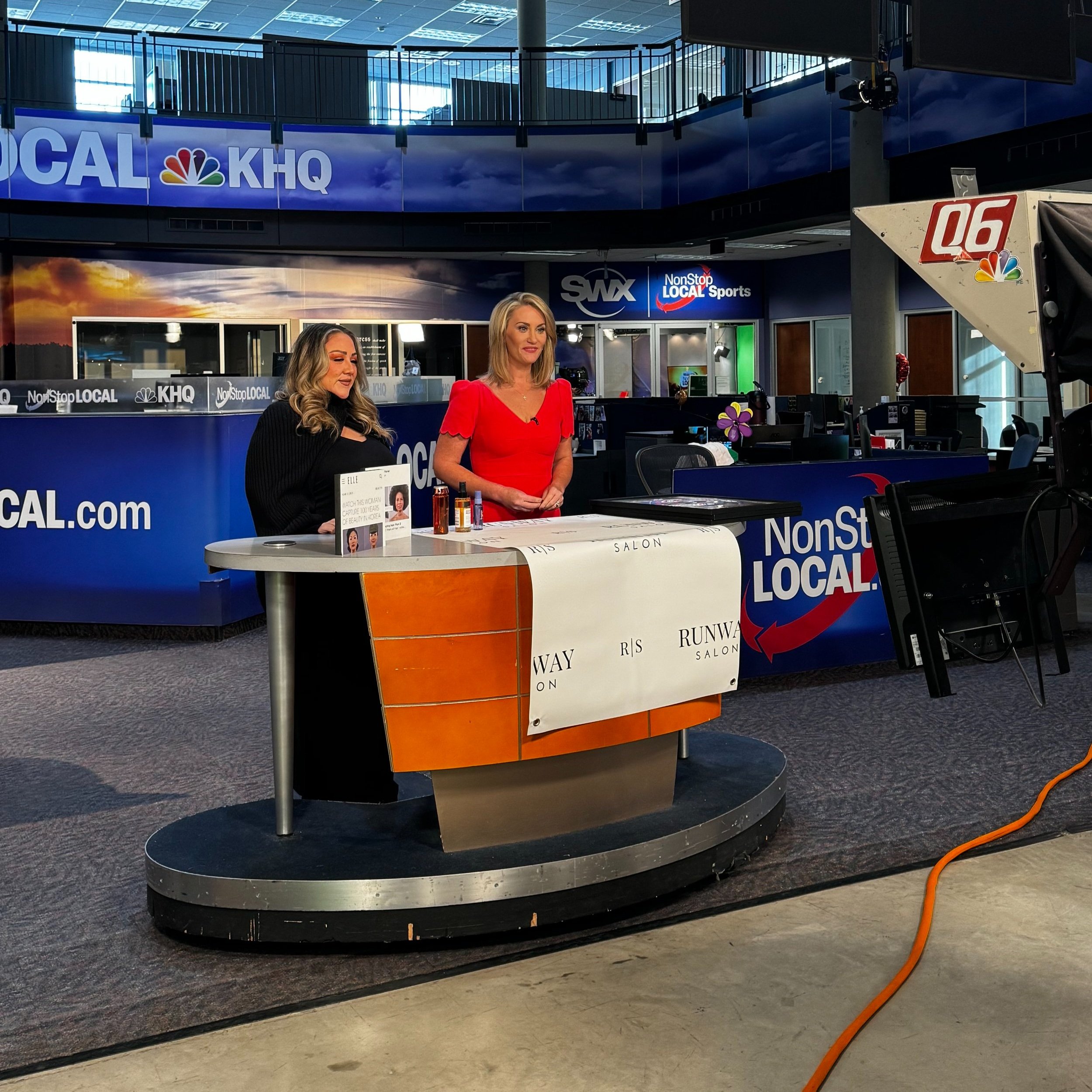 Two women are standing behind a news set, with cameras and news station signs visible in the background. One woman is wearing a red dress, and the other woman is dressed in black. The setting appears to be a television studio.