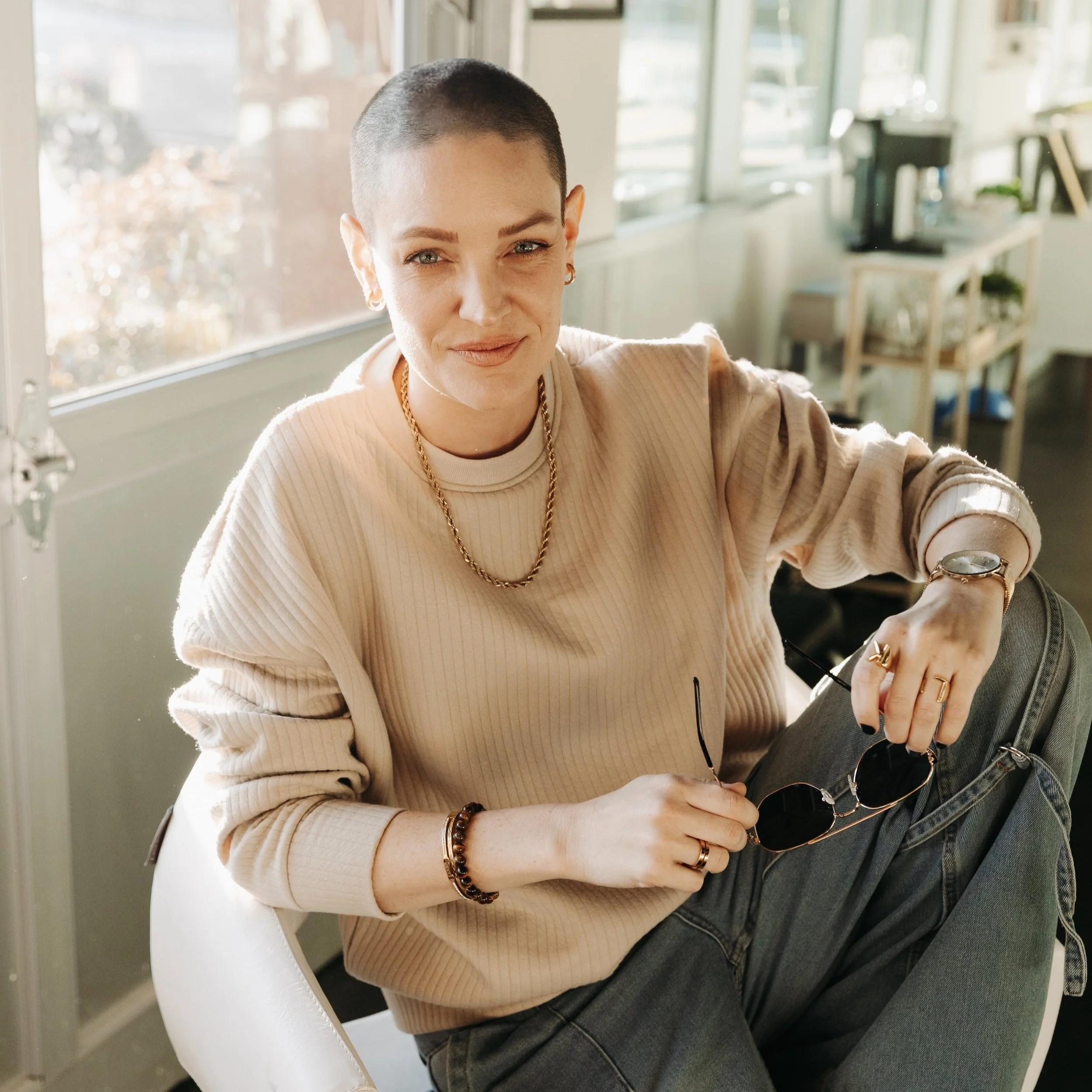 A woman with a shaved head sitting indoors, smiling at the camera, holding a pair of sunglasses in one hand, wearing a beige sweater, gold jewelry, and a wristwatch, with a casual environment in the background.