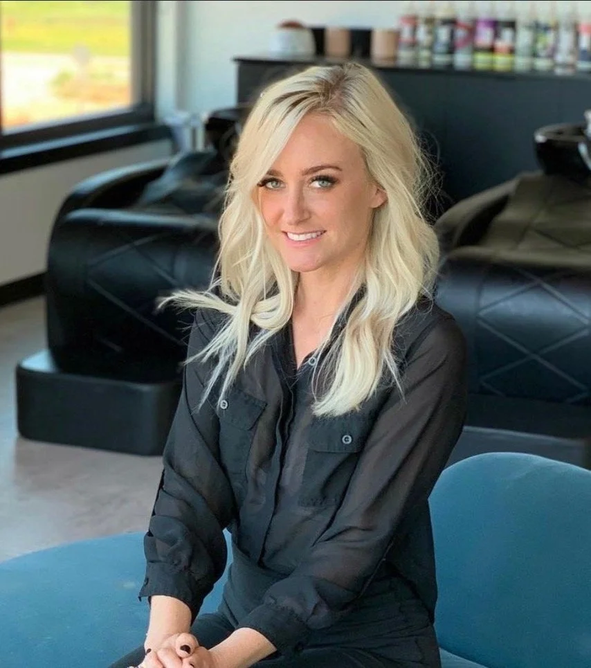 A blonde woman with wavy hair, wearing a black button-up shirt, smiling and sitting in a salon or hair studio. There are salon chairs and a large window in the background.
