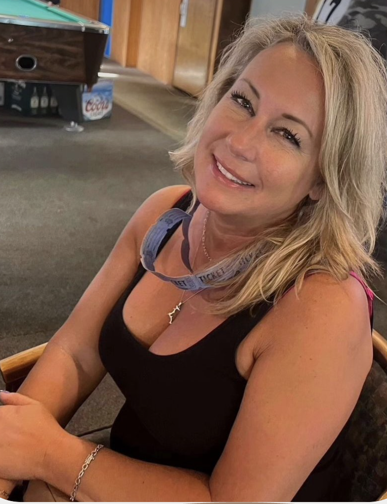 A woman with blonde hair smiling, wearing a black sleeveless top, sitting indoors with a fan and wooden furniture in the background.