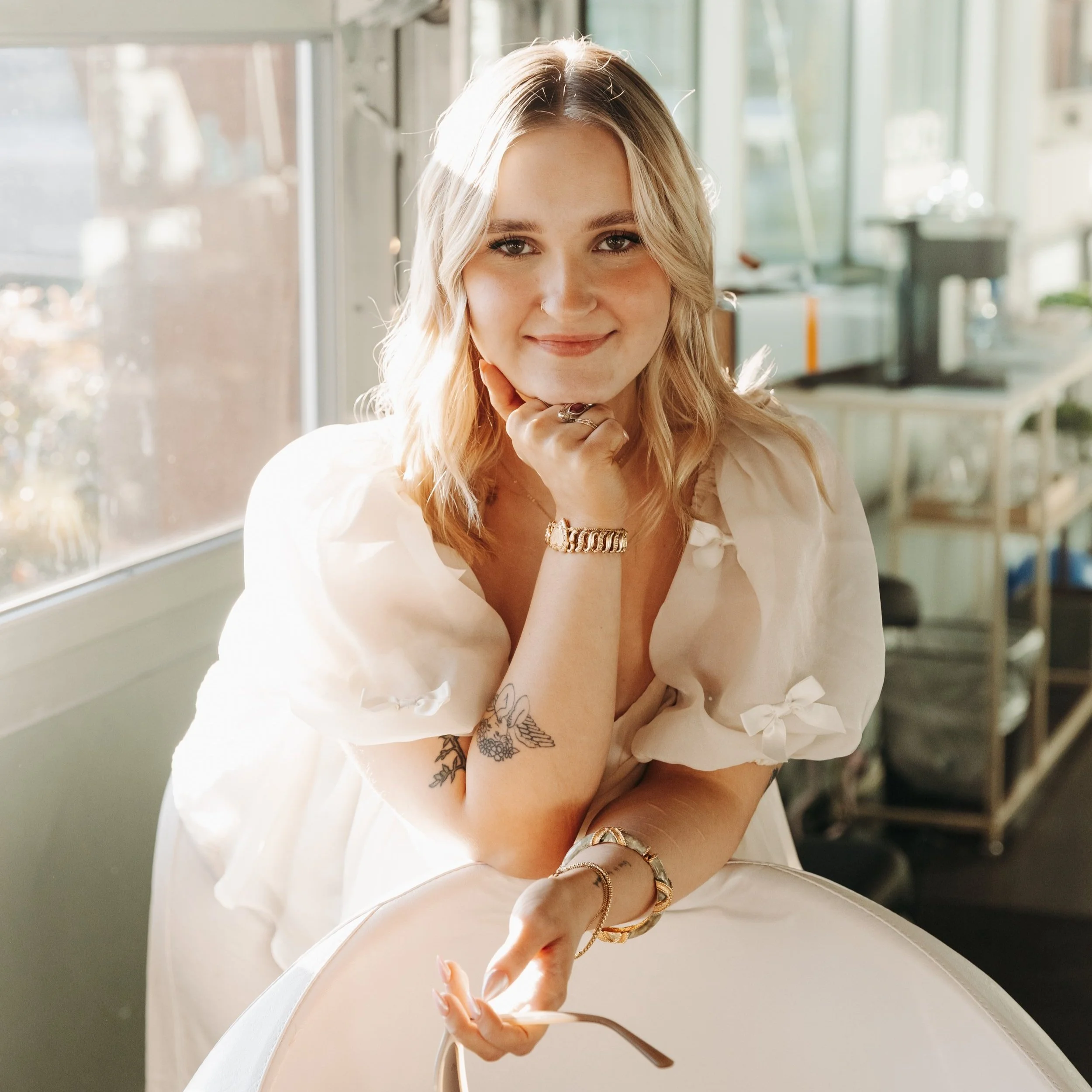 A young woman with blonde hair, wearing a white dress with tied sleeves, sitting by a window, smiling at the camera with her chin resting on her hand, in a bright indoor setting.