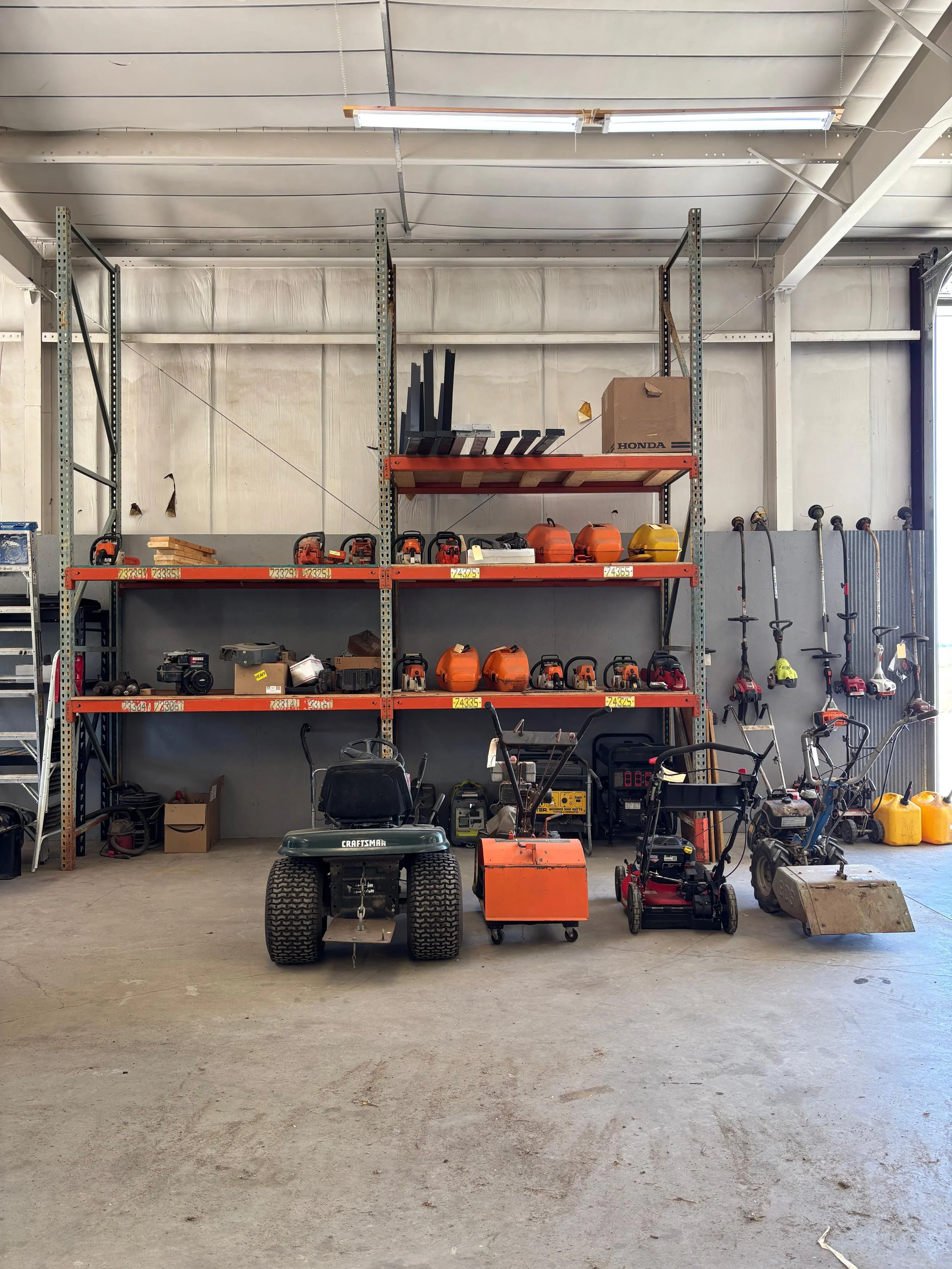 Organized workshop storage with lawn mowers, leaf blowers, chainsaws, and power tools on metal shelves.
