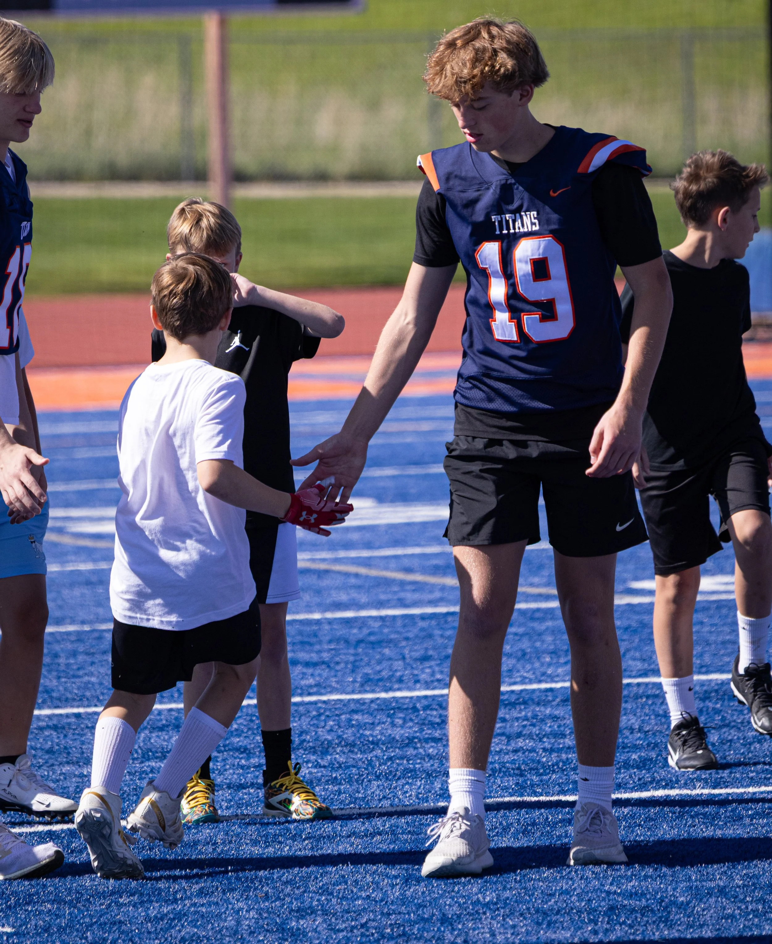 A group of children and a teenage boy playing on a blue sports field, with the teenage boy handing a red object to a young boy wearing a white shirt.