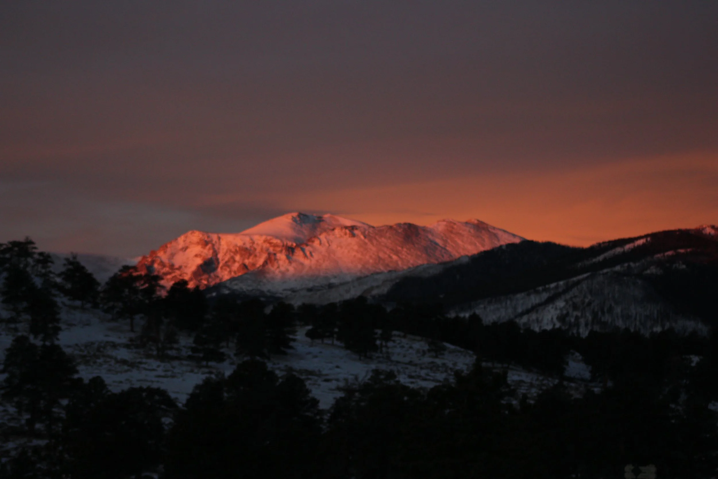 Snow-covered mountain at sunset, with orange-pink glow on its peak, dark silhouette of trees and rolling hills in foreground.
