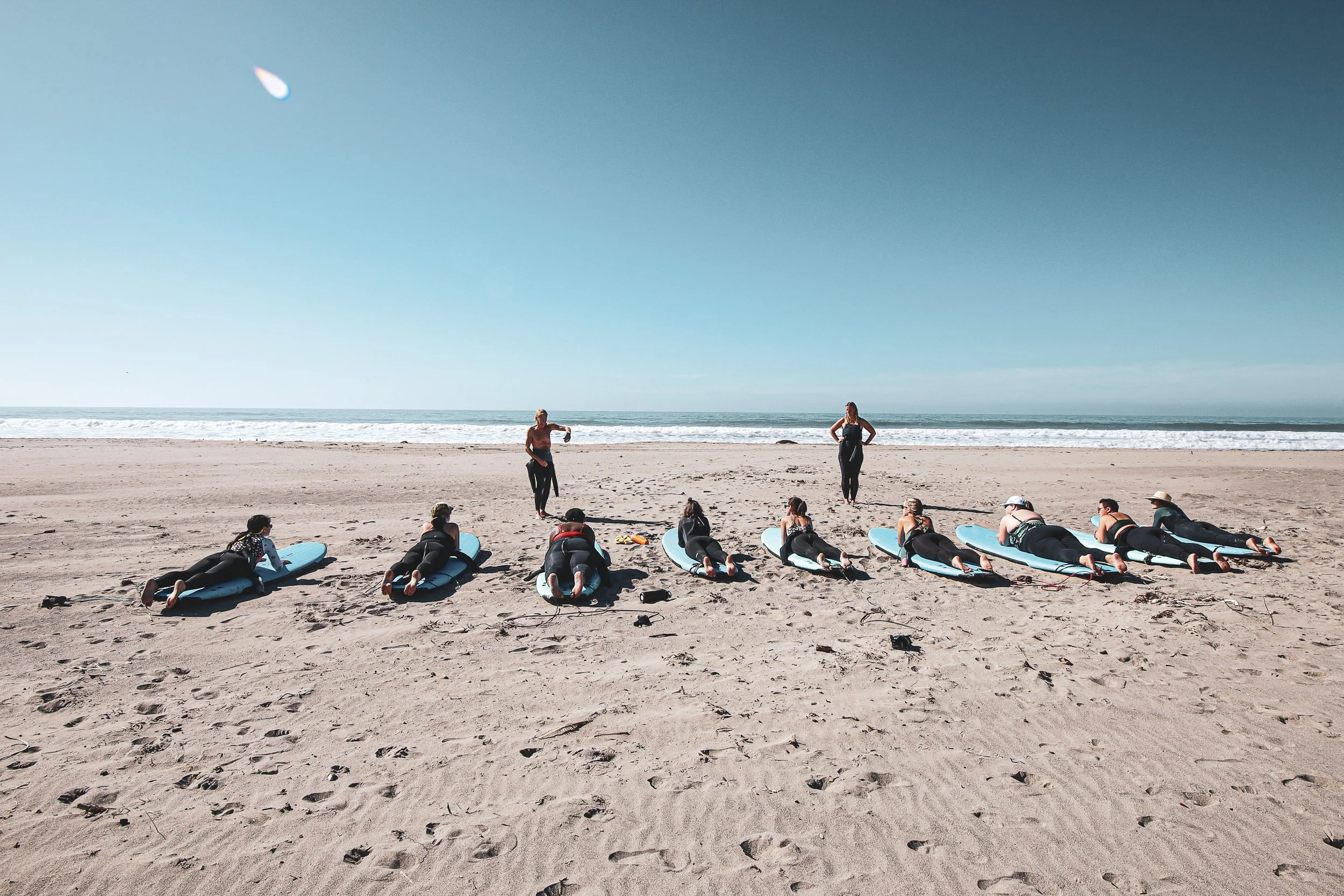 Group of people lying on surfboards on the beach during a surfing lesson, with instructors standing nearby, ocean and sky in the background.