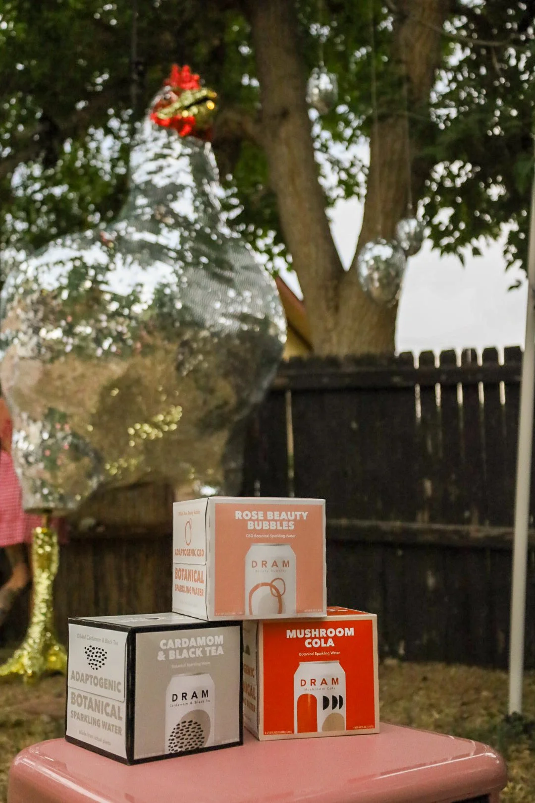 Three boxes of botanical sparkling water on a pink surface outdoors, with a tree and a black wooden fence in the background.