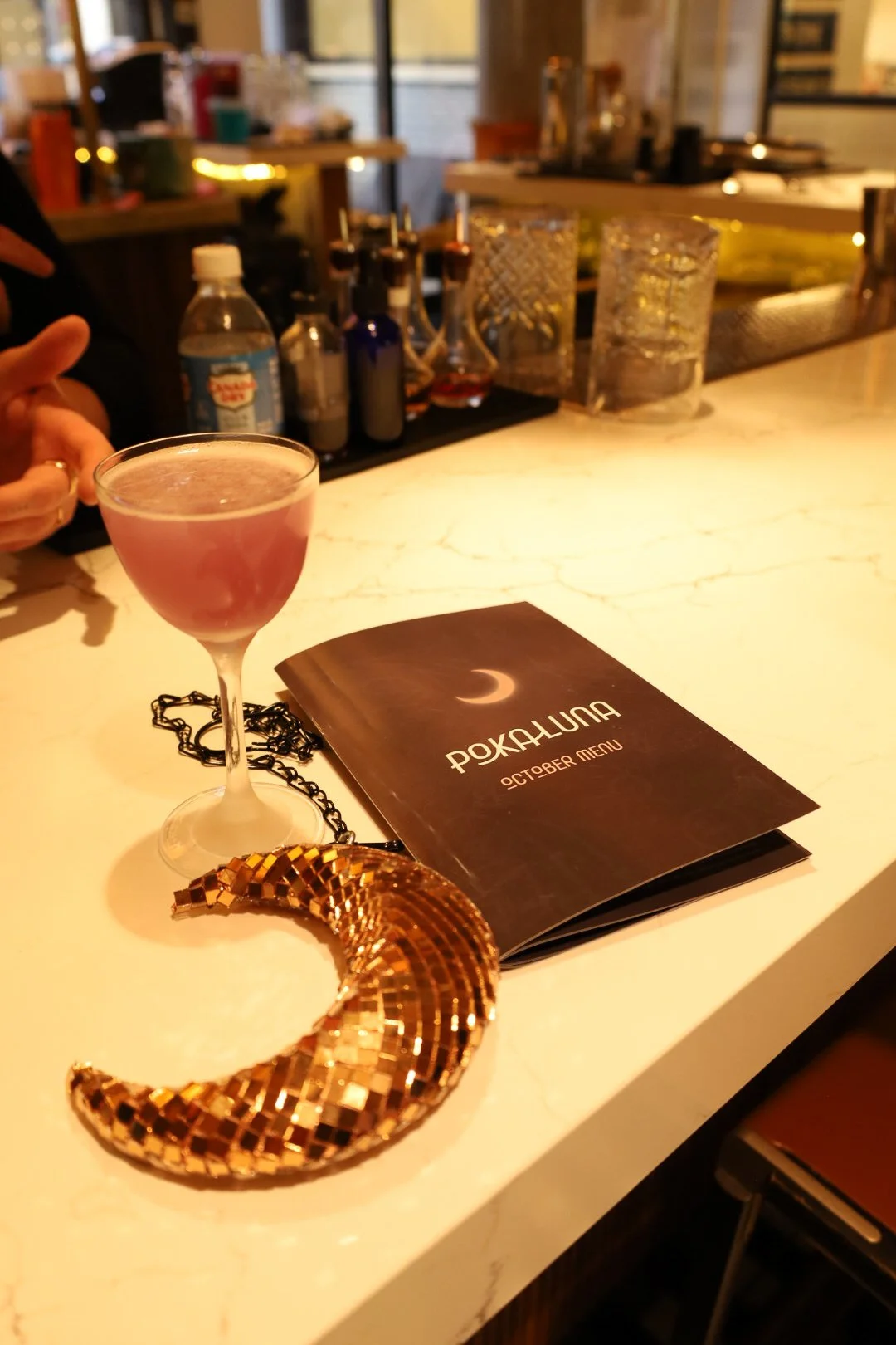 A cocktail glass with a pink drink, a folded October menu, a gold crescent-shaped art piece, and a gold bangle on a white marble bar counter, with a blurred background of bottles and glasses in a bar or restaurant setting.
