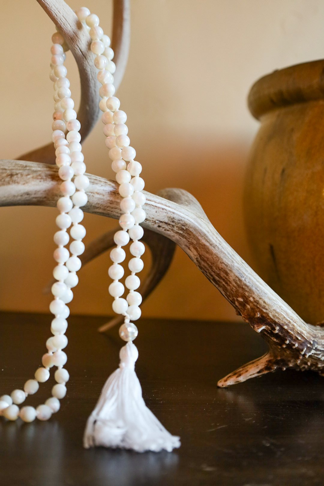 A white beaded mala necklace with a tassel hangs from a decorative antler against a warm background, with a brown jar in the background.