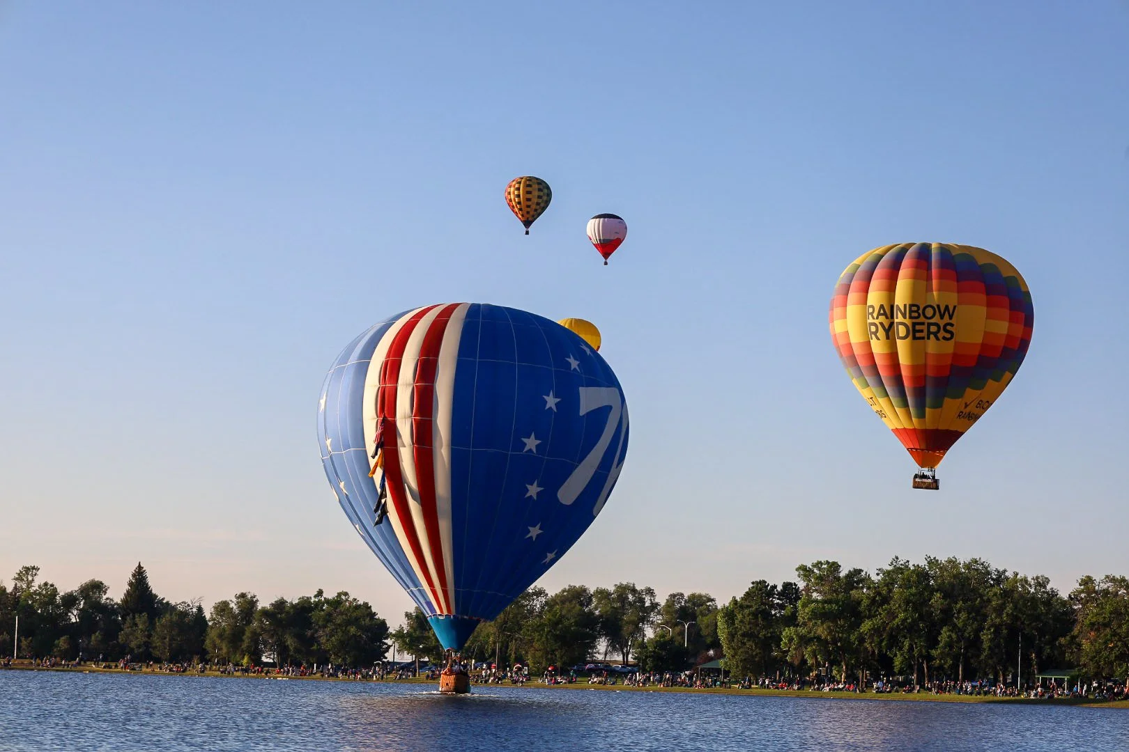 Several colorful hot air balloons flying over a body of water during daytime, with trees and a crowd of people on the shoreline.