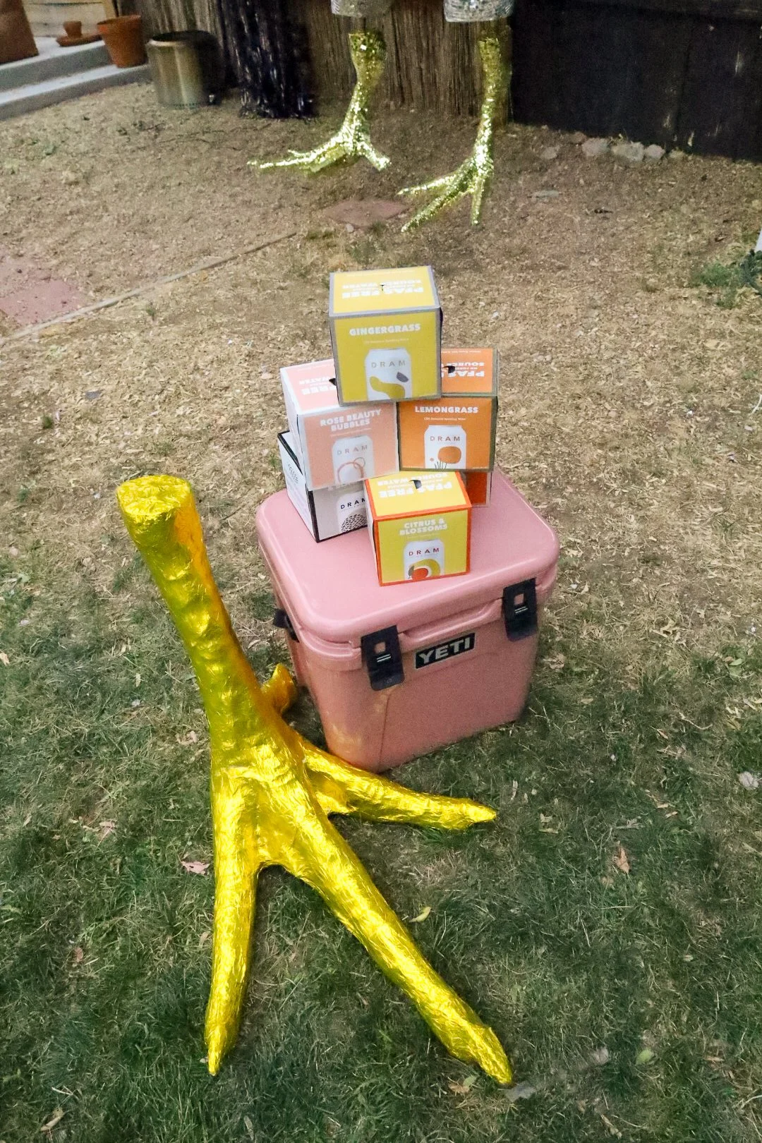 Decorative garden scene with a pink Yeti cooler, small colorful boxes of DRAM sparkling water stacked on top, metallic tree branch decorations, and a large yellow star-shaped sculpture.