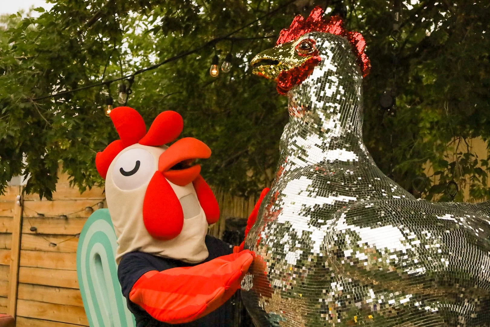A person in a chicken costume with a large chicken head and red comb, wearing orange gloves, stands next to a large metallic disco chicken sculpture covered in small reflective tiles, outdoors with trees and string lights in the background.