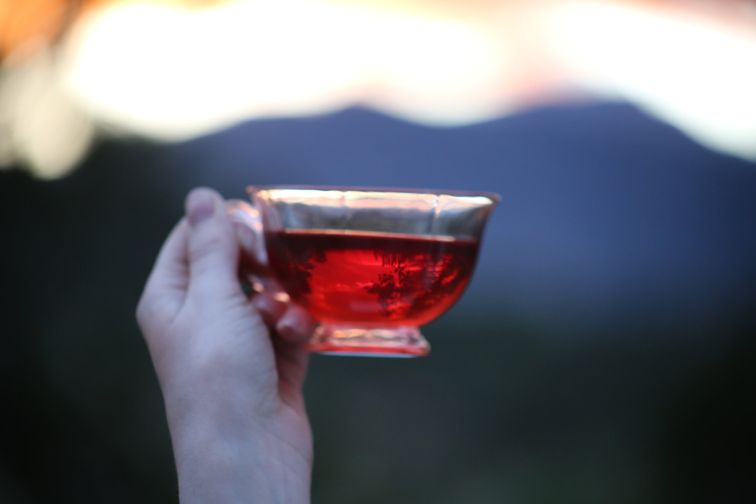 Person holding a glass cup filled with red tea, outdoors during sunset or evening, with a blurred mountain landscape in the background.